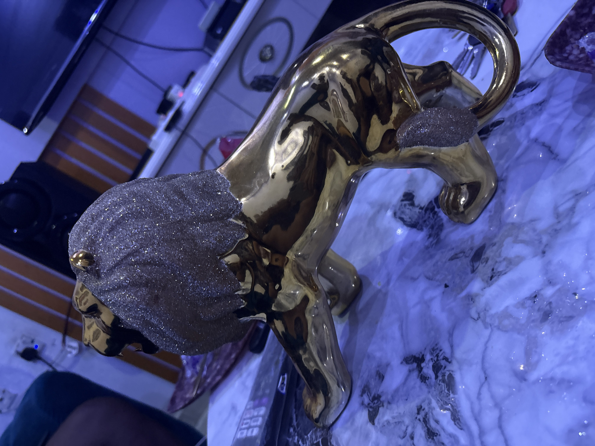 A golden lion figurine, adorned with glitter on its mane, sits on a marble-like surface under blue-tinted lighting. The figurine is positioned on its side, with its head and front paws slightly elevated. In the background, blurred electronic devices, including a speaker and a television, are visible. The scene appears to be indoors, likely in a domestic setting within the city of Obosi, Nigeria.