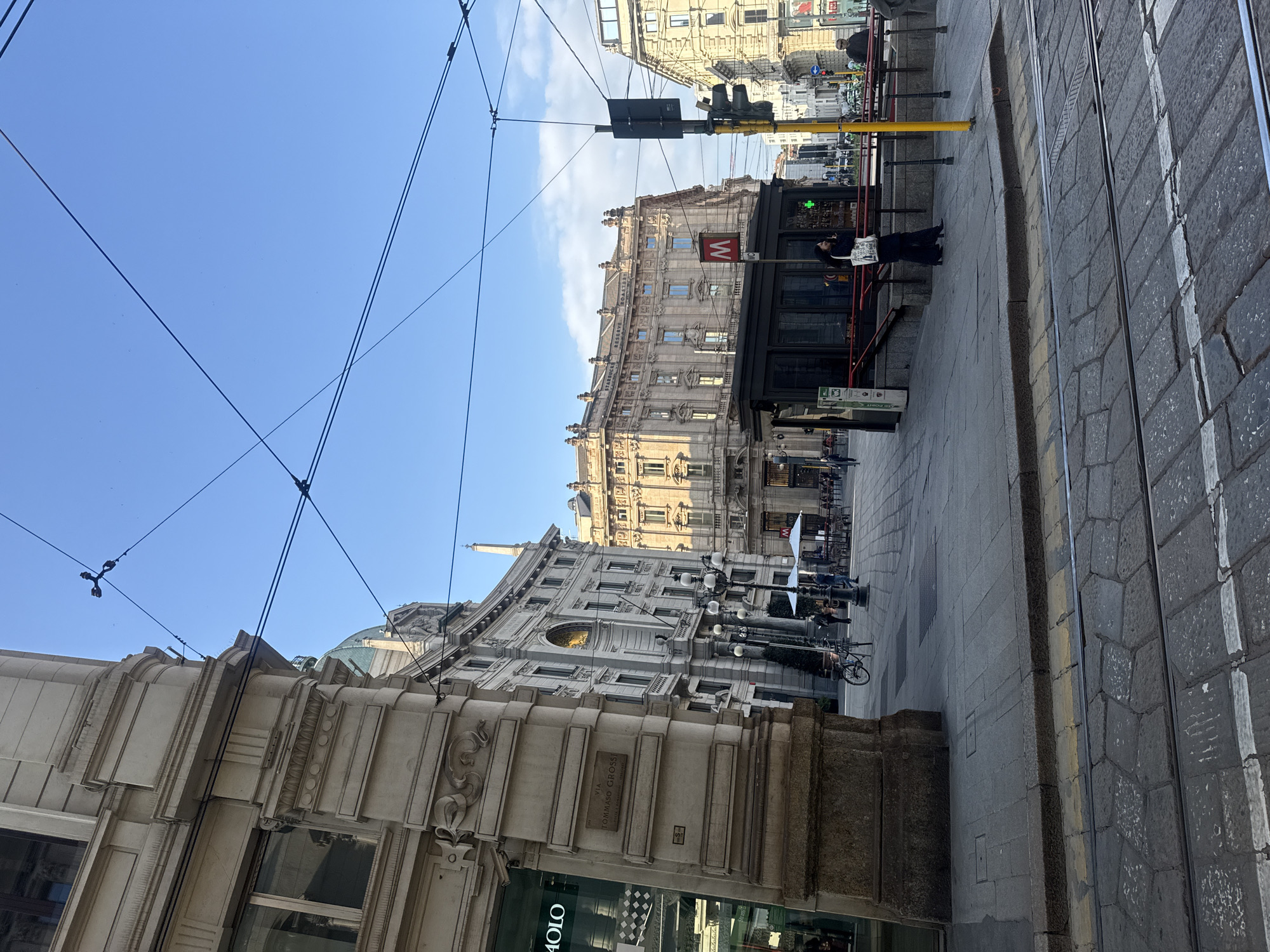 This is a street scene in Milan, Italy. The image is taken at street level, showing buildings with ornate facades. A Metro station is visible, marked with a large red 'M'. Pedestrians are present, including a woman in dark clothing standing next to the Metro station. Tram tracks run along the side of the street. Overhead electrical wires are visible against a blue sky. A street sign for Via Tommaso Grossi is visible on one of the buildings.