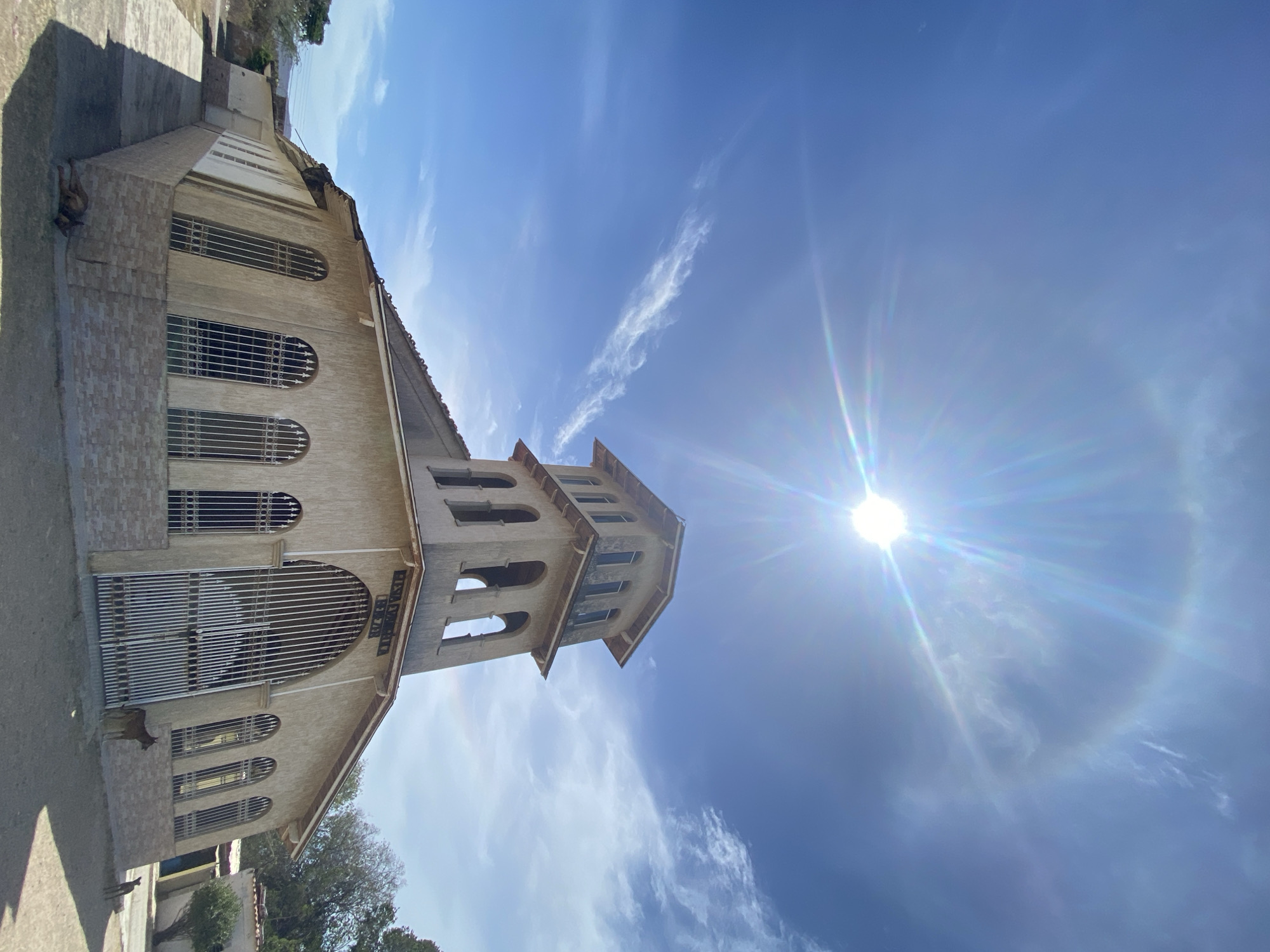 The image shows the Iglesia El Cristo del Buen Viaje in Juan Griego, Venezuela, viewed from a low angle looking up. The church is a beige building with arched windows and a bell tower. The sky is bright blue with some clouds and the sun is visible.