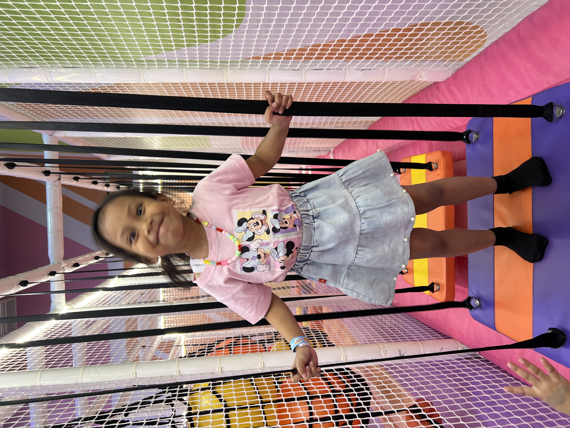 In Kecamatan Pati, Indonesia, a young girl stands in a colorful indoor playground. She is wearing a pink t-shirt with a Mickey Mouse graphic, a denim skirt, and black socks. A beaded necklace adorns her neck, and a blue wristband is on her left arm. The girl is smiling and looking at the camera. She is standing on a suspended platform, with netting and poles surrounding her. The playground features bright colors like pink, purple, orange, and green.