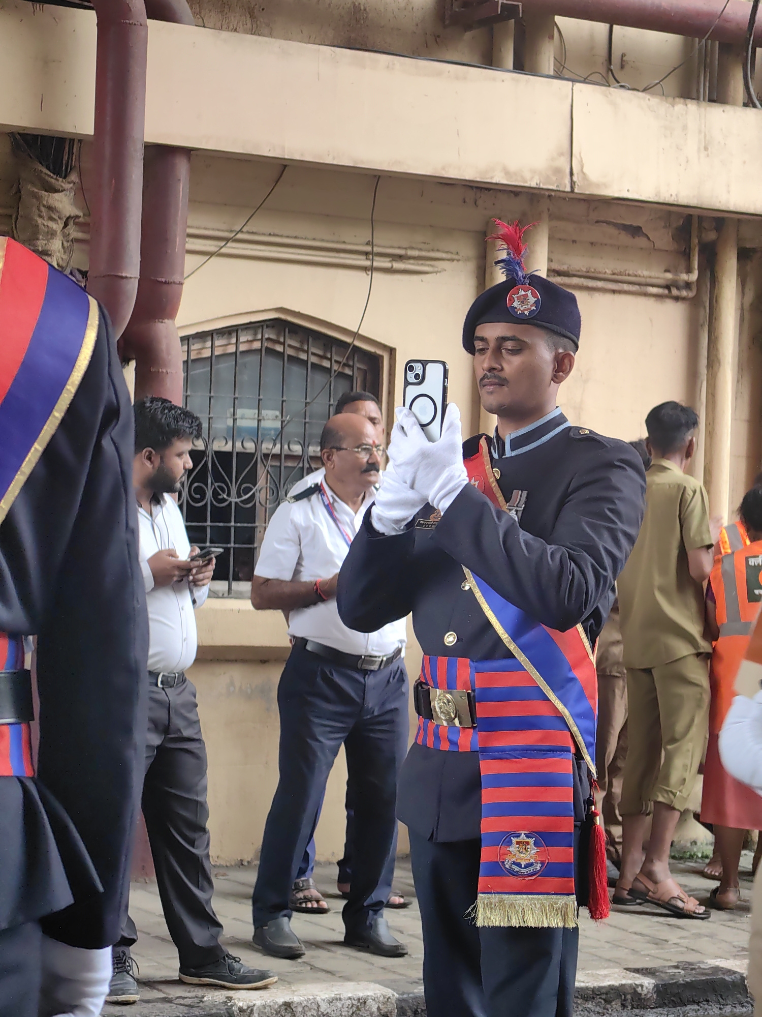 In the city of Mumbai, India, a young man in a dark blue military uniform, complete with a beret adorned with a red and blue feather, a sash of red and blue stripes, and a medal, is captured mid-action. He is holding a white smartphone in both hands, seemingly taking a photo or recording a video. He wears white gloves.
Several other individuals are visible in the background. To his left, a man in a white collared shirt and dark pants looks down at his own phone. Further left, another person in a dark uniform with red and blue trim is partially visible. Behind the main subject, a man in a white shirt and dark pants stands attentively, and another man wears a light brown uniform. To the far right, a person in an orange safety vest is partially seen.
The backdrop is an exterior wall of a building, appearing somewhat aged, with visible pipes running along it. A barred window is also noticeable. The ground is paved with cobblestones and some concrete slabs. The overall scene suggests an outdoor event or gathering, possibly a parade or ceremonial occasion.