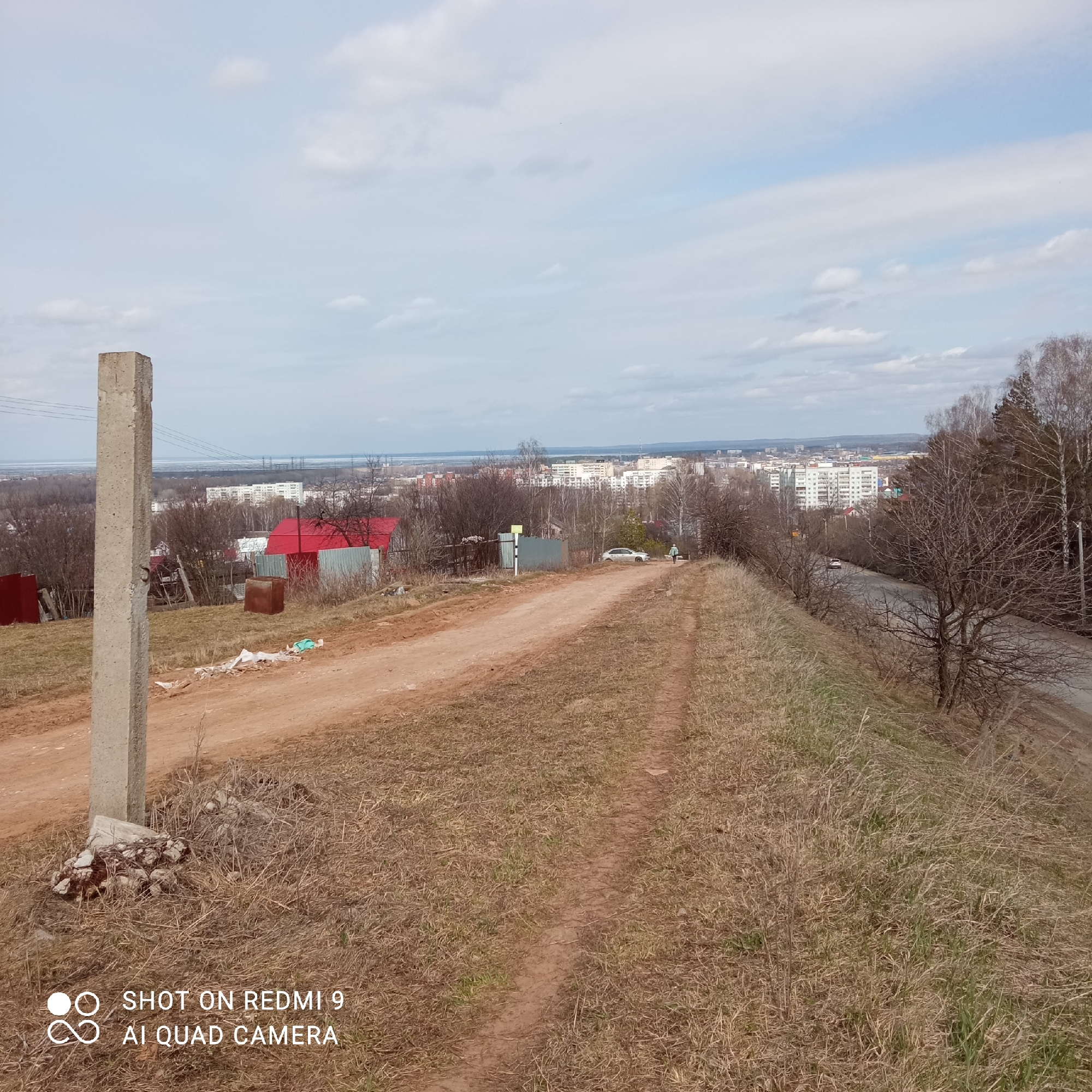 The image captures a panoramic outdoor scene from an elevated vantage point, looking down upon a semi-urban landscape in Chaikovsky, Russia, during what appears to be late winter or early spring.
In the foreground, a tall, rectangular concrete post stands prominently on the left, with some debris scattered at its base. A wide, well-trodden dirt path dominates the center-left, with a narrower foot trail diverging towards the right side. The ground is largely covered in dry, light brown grass, interspersed with some areas of new green growth.
The mid-ground reveals a mix of natural and man-made elements. Numerous bare trees and bushes are scattered across the slopes. Several small structures are visible, including a building with a bright red roof and various corrugated metal fences (red, grey, and green). A white car is parked off the dirt path, near a small yellow-green sign. Further into the mid-ground, a paved road can be seen, with a second, lighter-colored car traveling along it. A single person, dressed in a light-colored top and dark pants, is walking away from the camera along the narrower dirt path on the right.
The background showcases a sprawling town or city, characterized by numerous multi-story residential buildings. Beyond the urban area, a wide body of water, likely the Kama River, extends to the horizon under a hazy sky, with power lines or transmission towers visible in the far distance. The sky above is partly cloudy, with some patches of blue visible. The lighting indicates it is daytime.
Text watermarks in the bottom-left corner read "SHOT ON REDMI 9" and "AI QUAD CAMERA".
