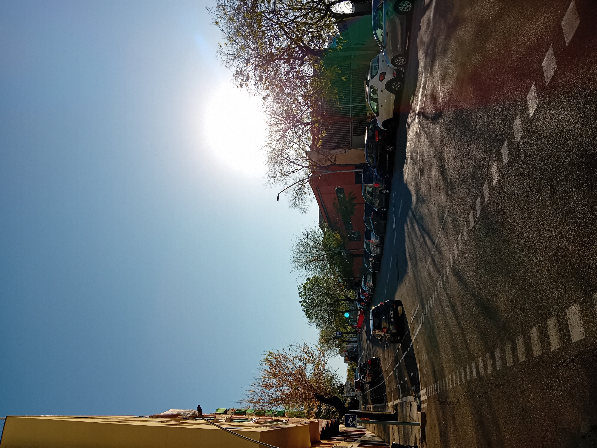 The image depicts a street scene in Barcelona, Spain. The perspective is tilted, showing a clear blue sky with a bright sun. Buildings with yellow and red facades line the left side of the street. A row of parked cars is visible on the right side. Trees with sparse foliage are present on both sides of the street, casting shadows on the asphalt. A black car is driving down the street. There are white rectangular markings on the road. A bicycle lane symbol is visible near the yellow building. A street lamp is visible near the red building.