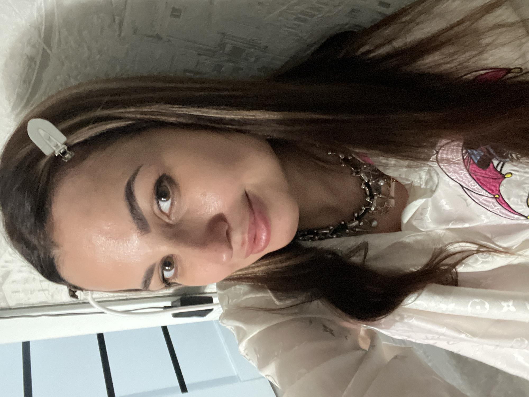 A woman with brown hair and a white clip is facing the camera. She is wearing a necklace and a white top with a pink and yellow butterfly design. The background is a white wall and a blue and white surface. The location is Unknown, Unknown.