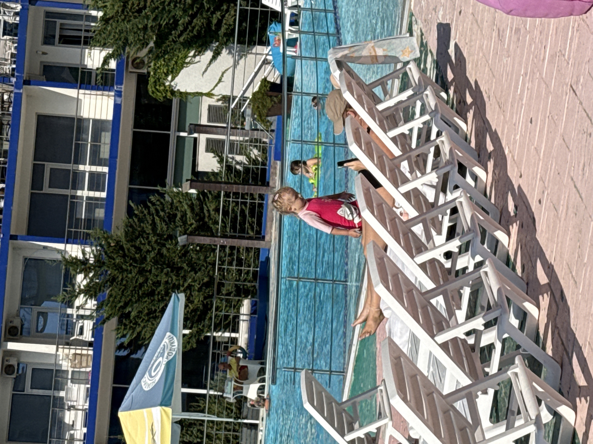 In Sevastopol, Ukraine, a bright, sunny day is depicted at a resort, likely near a beach or pool. In the foreground, several white plastic lounge chairs are lined up on a reddish-brown tiled patio. The chairs are mostly empty, with one person partially visible lying on a chair, wearing a hat and holding a phone. A young child in a pink swimsuit stands near the chairs, looking towards the pool.
In the background, a large, bright blue swimming pool is active with people. A few individuals are swimming or relaxing in the water. Some are on inflatable toys, including a yellow one with a child on it. A lifeguard, wearing a yellow shirt and sitting on a white chair, is visible near the edge of the pool. Behind the pool area, a modern multi-story building with balconies and windows is visible, surrounded by lush green trees and ivy. A yellow and blue umbrella is set up near the pool, offering shade. The overall atmosphere suggests a leisurely vacation setting.