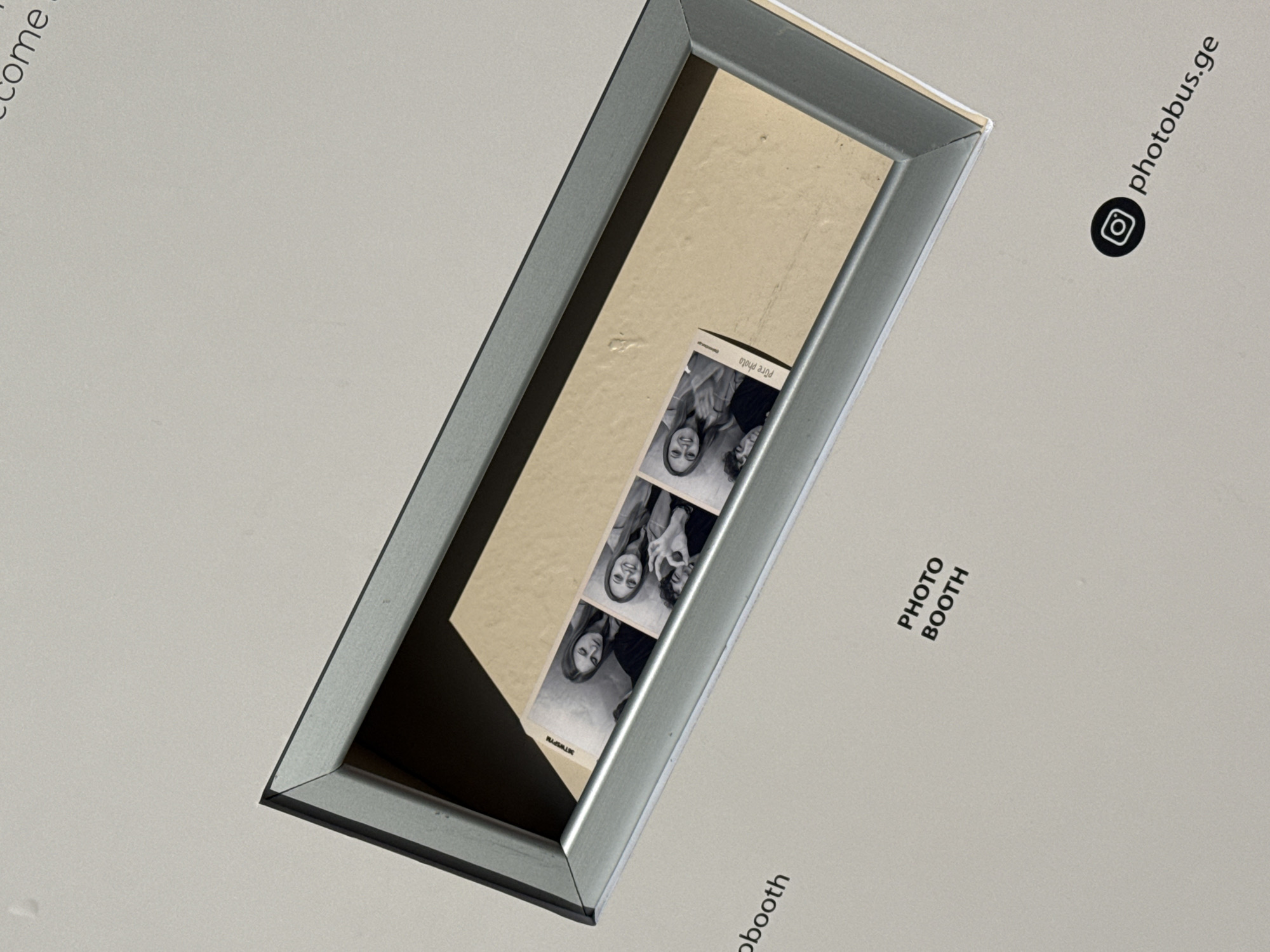 The image presents an angled, overhead view of what appears to be a photo booth display or output slot, situated on a clean, light grey surface, likely indoors. A prominent rectangular opening, framed by sleek silver metallic edges, is seen receding into a light brown-beige interior.
Inside this framed opening, a black and white photo strip is casually placed, slightly askew. The strip features four individual frames, each capturing two people—a man and a woman—posing. Their expressions are cheerful and playful, with visible smiles and an instance of a "rock on" hand gesture. Faint text "Pure photo" is discernible at the top of the photo strip.
The surrounding light grey surface carries several textual and graphical elements. In the upper right section, oriented diagonally, the text "photobus.ge" is displayed alongside a black Instagram logo. Directly below this, also diagonally, the words "PHOTO BOOTH" are printed. A partial word, "come," is visible in the upper left corner of the frame, hinting at a larger welcome message.
The overall scene suggests a modern and interactive service, likely a photobooth, found in Batumi, Georgia, based on the provided context and visible branding. The aesthetic is clean and contemporary, emphasizing the fun and personal memento of photo booth strips.