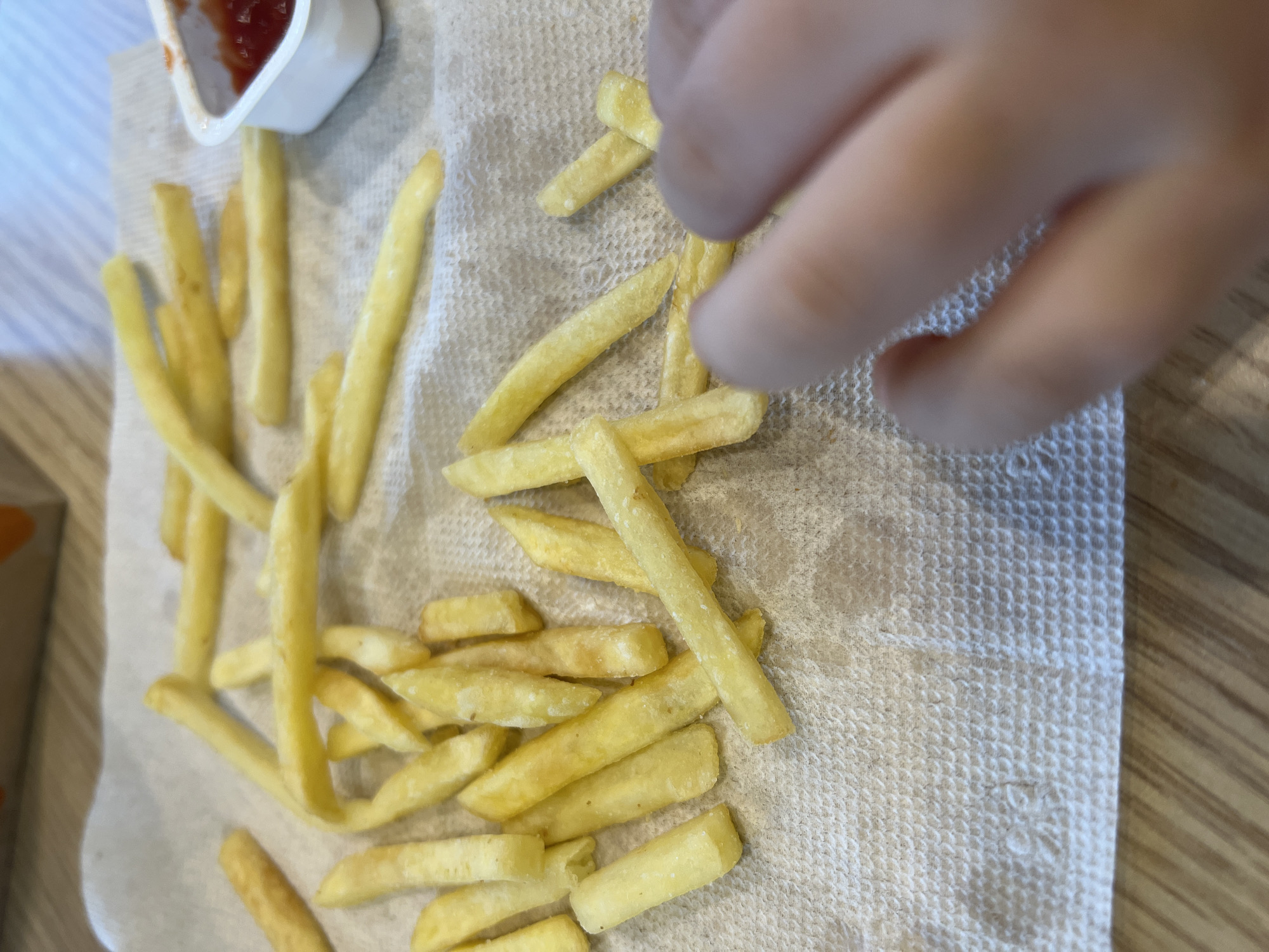 A close-up, overhead shot shows a pile of french fries spread across a white paper napkin on a wooden table. A small white container of ketchup is visible to the right, filled with red sauce. A child's hand with lightly tanned skin is reaching for the fries from the right side of the frame. The background is blurred but suggests an indoor setting. No specific location in Yekaterinburg, Russia, can be determined from the image.