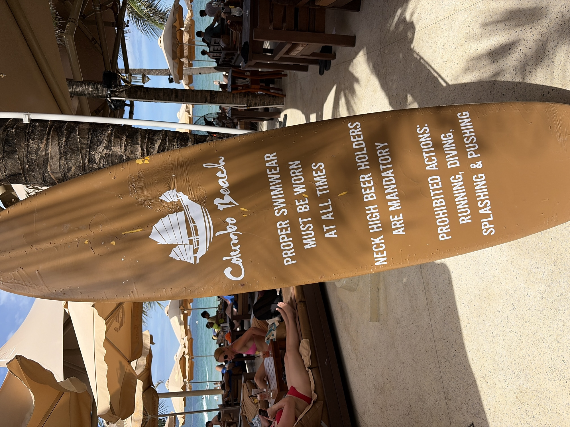 A surfboard-shaped sign stands in the foreground, displaying rules for Colombo Beach in Đà Nẵng, Vietnam. The sign is a light brown color with white text. It states: "COLOMBO BEACH", "PROPER SWIMWEAR MUST BE WORN AT ALL TIMES", "NECK HIGH BEER HOLDERS ARE MANDATORY", and "PROHIBITED ACTIONS: RUNNING, DIVING, SPLASHING & PUSHING".
In the background, several beach umbrellas provide shade for lounge chairs, where people are relaxing. The ocean is visible in the distance, and palm trees are interspersed throughout the scene. The ground is paved with speckled tiles. The overall setting is a beach resort or restaurant.