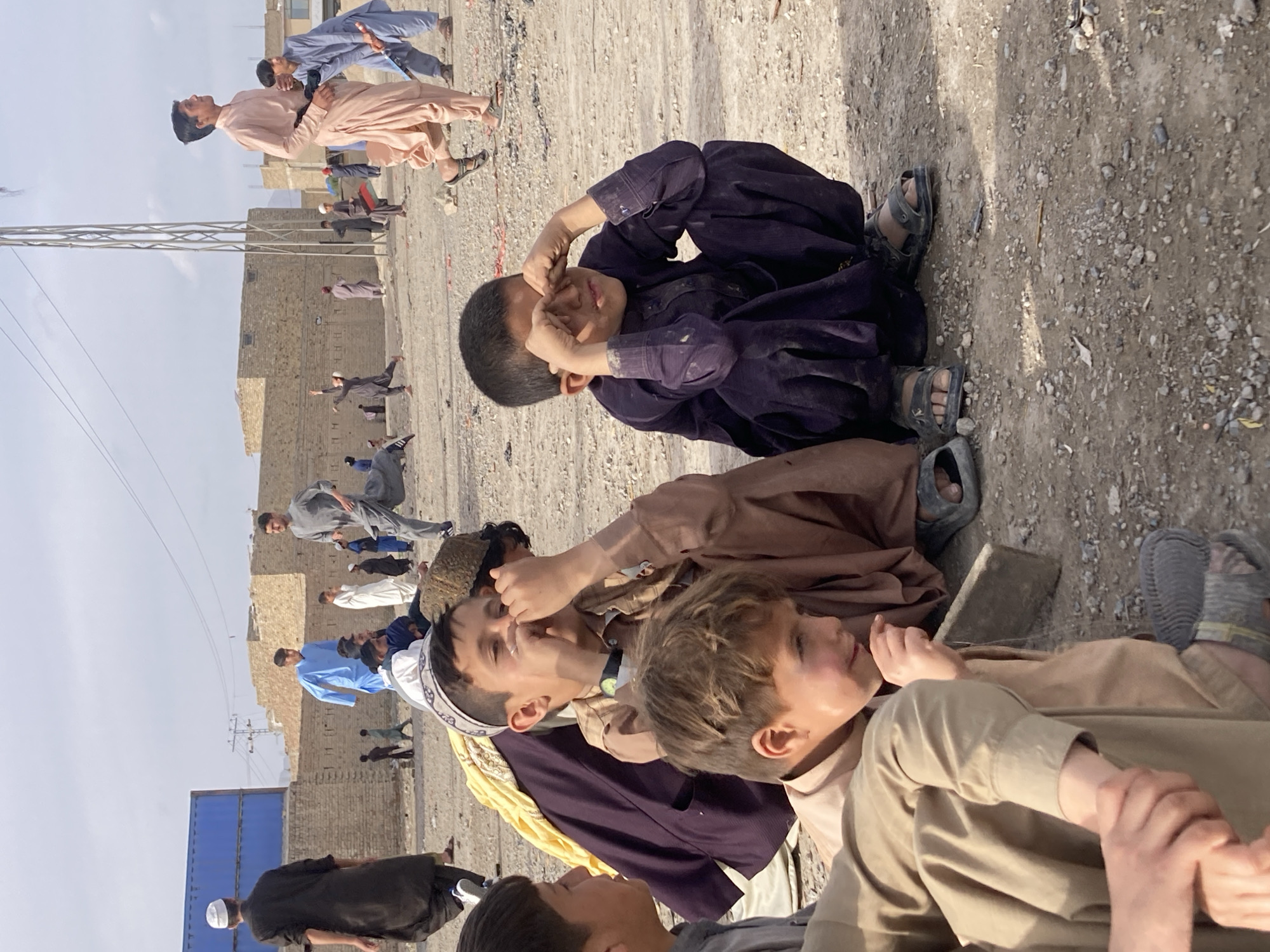 This image depicts an outdoor, unpaved open area in Quetta City Tehsil, Pakistan, populated by multiple males of varying ages, primarily children and young adults. The ground is dry, light brown earth covered with small stones and scattered debris.
In the immediate foreground, three young boys are positioned. The central boy, wearing a light brown shalwar kameez and a white embroidered skullcap (topi), sits with his hands covering his nose and mouth, his eyes partially closed. To his right, another boy in a dark purple shalwar kameez crouches, rubbing his eyes with both hands. A third boy, wearing a light brown shalwar kameez, is partially visible on the far left, looking to the right.
In the midground and background, numerous other males are engaged in various activities. A young man in a light peach-colored shalwar kameez walks briskly from right to left, carrying a dark object resembling a toy weapon or stick. Another young man in a grey shalwar kameez runs from left to right. Several other individuals, dressed in various colored shalwar kameez (including blue, white, and black), are walking, standing, or running further back.
The background features low-rise buildings constructed from light-colored bricks or mud bricks, some with flat roofs. On the far left, a large blue corrugated metal shipping container is visible. A tall, open-lattice metal utility tower stands prominently on the right side of the frame. The sky is a pale, hazy blue.