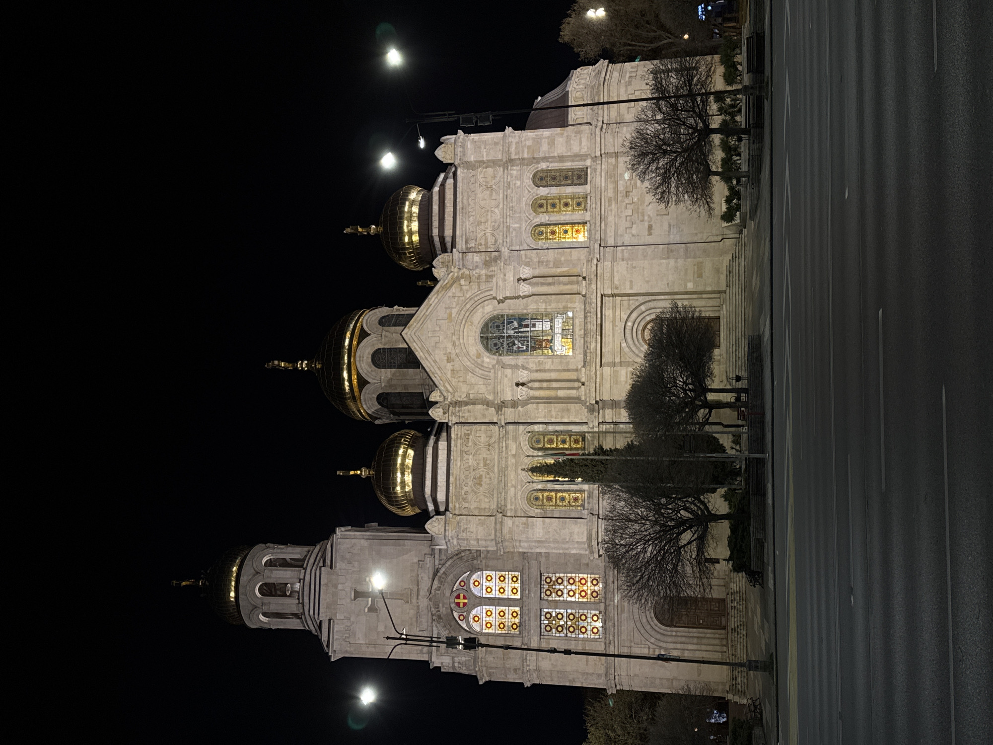 The image captures the Cathedral of the Assumption of the Virgin in Varna, Bulgaria, at night. The cathedral is illuminated, highlighting its architectural details, including its golden domes and stained-glass windows. A street runs alongside the cathedral, and there are bare trees planted nearby. The background is dark, emphasizing the illuminated building. There are street lights visible, and the street appears empty of people or vehicles.
