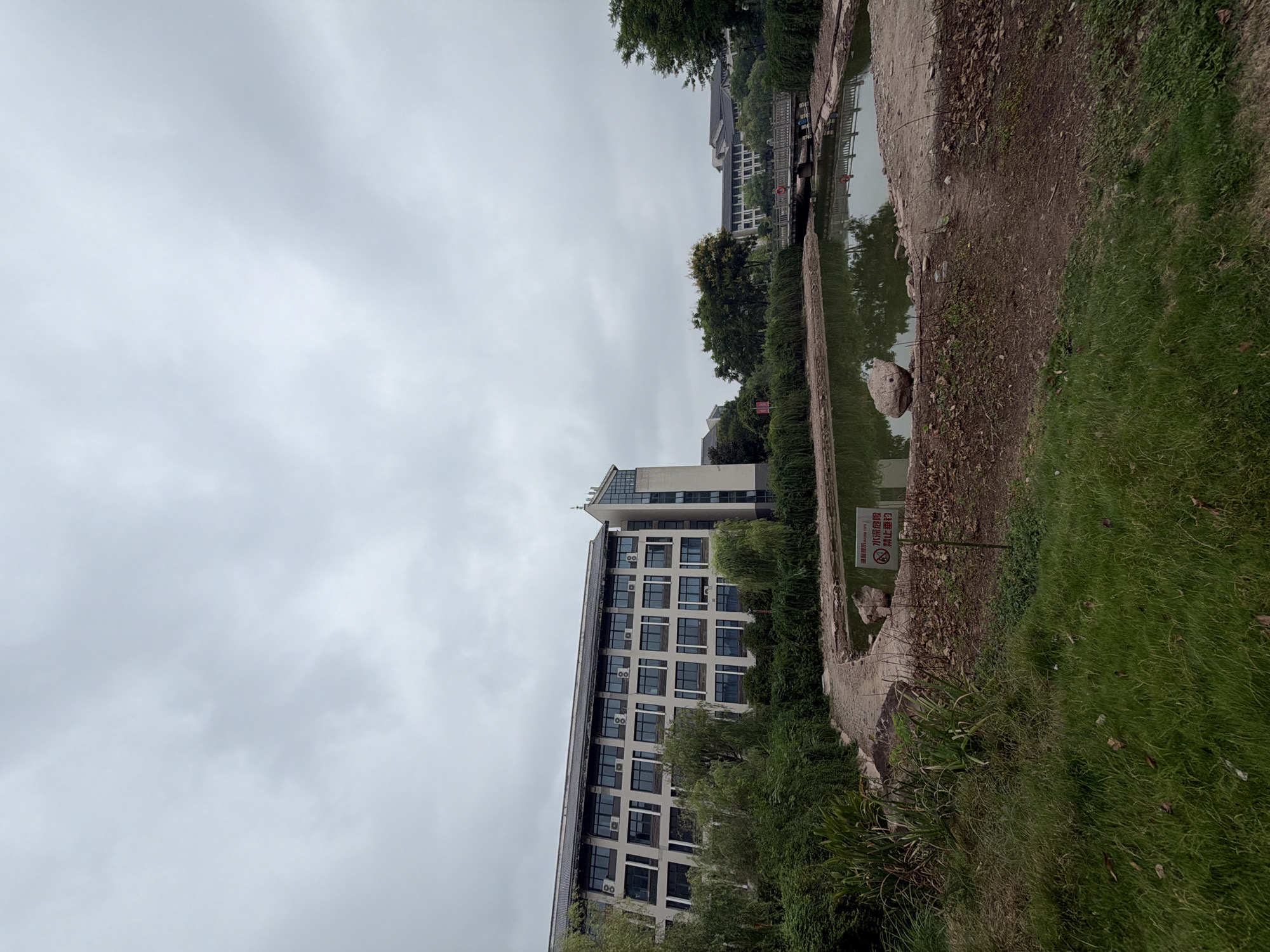 The image depicts an outdoor scene on a cloudy day, likely in Jiangjin, China, featuring a body of water resembling a pond or slow-moving river. In the background, a multi-story modern building with numerous windows and air conditioning units is visible, surrounded by lush green trees. A pathway made of stone or gravel curves along the edge of the water. On the right side of the frame, a bridge spans the water, with another building visible beyond it. A small sign is planted near the pond's edge, bearing Chinese text and a prohibitory symbol. The foreground is dominated by grass and some patches of bare earth. There are no people visible in the image.