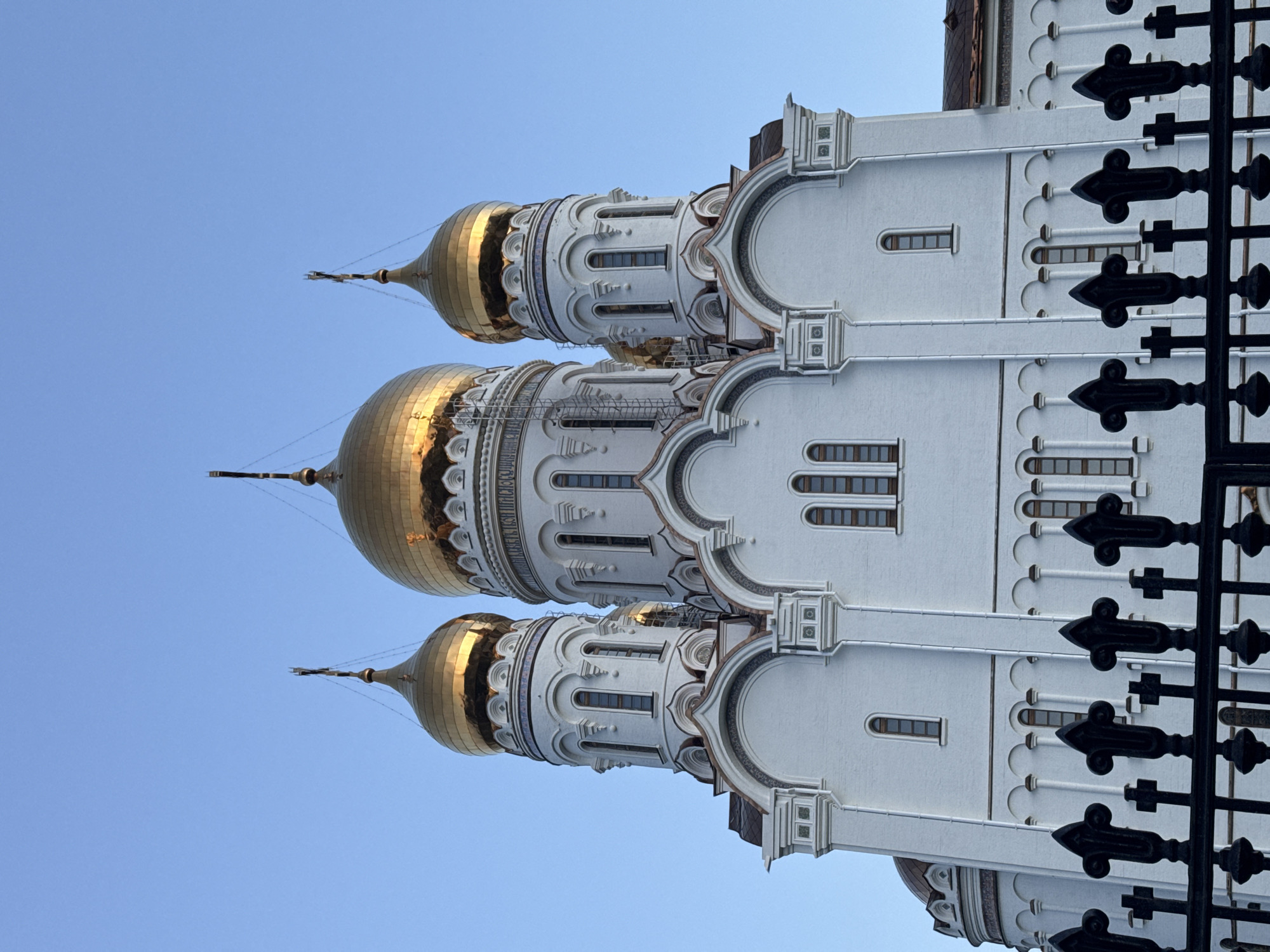 The image depicts a building with three golden domes against a clear blue sky. The domes appear to be part of a church or cathedral. The building is predominantly white with detailed architectural features and ornamentation. A wrought iron fence with decorative elements is visible in the foreground on the right. The image is taken from a low angle, emphasizing the height of the structure. The location in the city of Unknown, Unknown is not possible to determine.