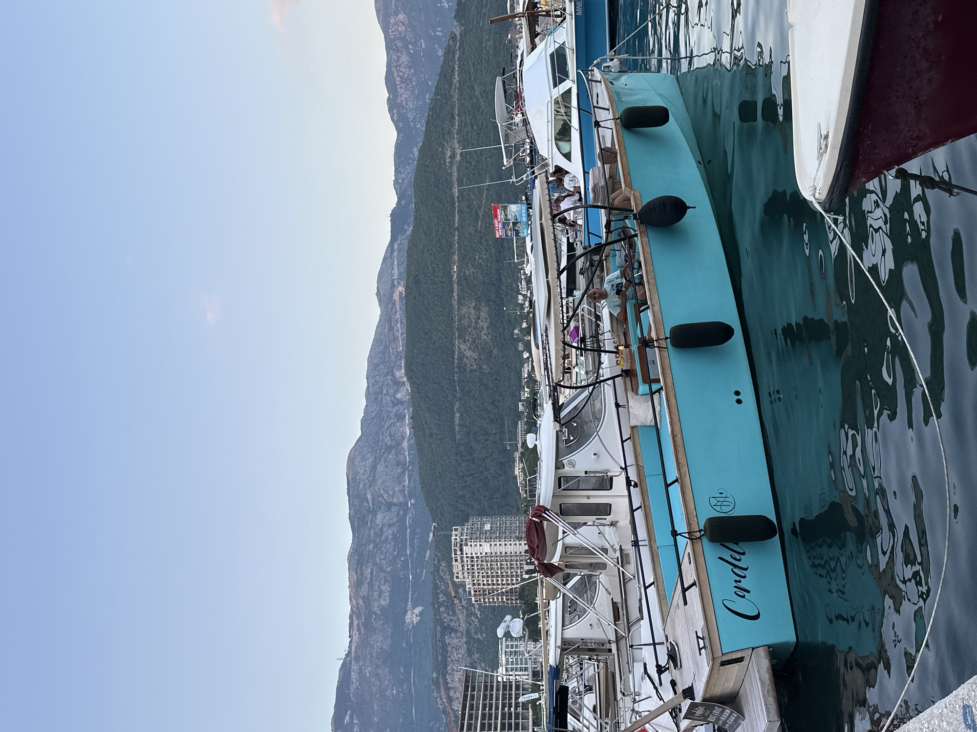 The image depicts a serene view of a marina in Budva, Montenegro, likely at dusk given the soft lighting. In the foreground, a bright turquoise boat with the name "Cordelia" emblazoned on its side is prominently featured. The boat has a wooden deck at the stern and a canopy with seating covered in light blue cushions. Several black fenders hang from its side. A man, wearing a light-colored t-shirt, is seated at the helm of the turquoise boat, appearing relaxed.
Behind the turquoise boat, other vessels are moored, including a white boat with a white cabin and canopy. In the mid-ground, several buildings are visible, including a large, modern, multi-story structure that appears to be under construction, with visible scaffolding and cranes. Further in the distance, lush green mountains rise, forming a dramatic backdrop against the pale blue sky. A winding road can be seen traversing the mountainside. A red and white sign with text, likely advertising boat rentals, is also visible on one of the boats. The water in the marina is calm, reflecting the boats and the sky, with visible ripples from the gentle movement of the water. A white rope is tautly stretched across the bottom foreground, connecting two points along the edge of the marina. The overall scene conveys a sense of tranquility and leisure within a coastal urban environment.