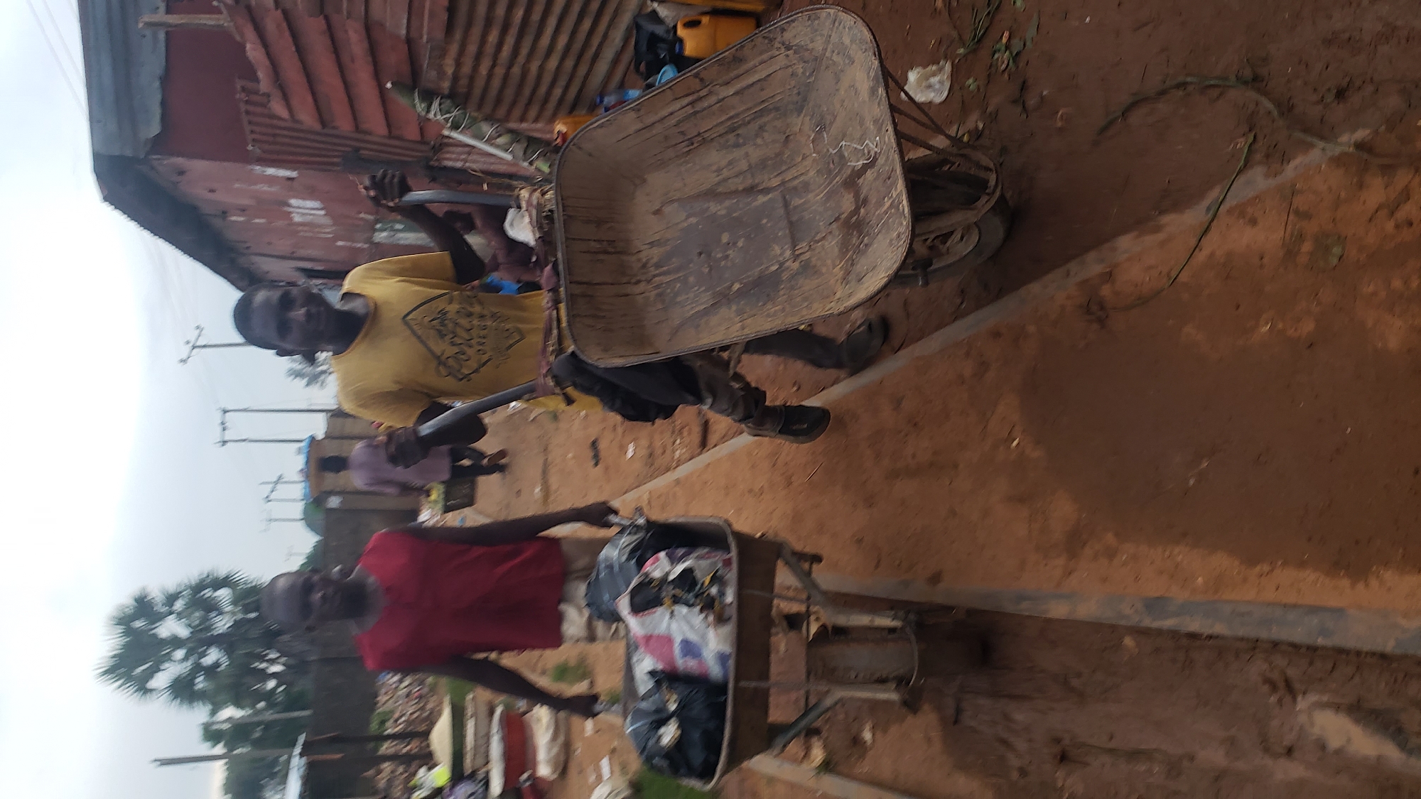 The image shows two young men pushing wheelbarrows in what appears to be an outdoor market in Kakuri Gwari, Nigeria. The ground is a mix of dirt and mud, with some debris scattered around. The sky is overcast, suggesting cloudy weather.
In the foreground, on the left, a man wearing a red sleeveless shirt is pushing a wheelbarrow loaded with black trash bags and some white and patterned bags. He is looking forward, focused on his task. To his right, another young man in a yellow t-shirt is pushing an empty, mud-caked wheelbarrow. He is looking towards the camera with a slight smile and appears to be holding a metal rod or pole connected to the wheelbarrow. His clothing includes a dark jacket or shirt draped over his shoulders and camouflage-patterned pants.
In the background, other individuals are visible, one in a light purple garment walking away from the camera, and others are partially obscured. The structures in the background seem to be makeshift market stalls with corrugated metal walls and roofs. One building on the right is red with visible signs of wear and tear. There are also some yellow jerrycans visible. The overall environment suggests a busy, working-class area.