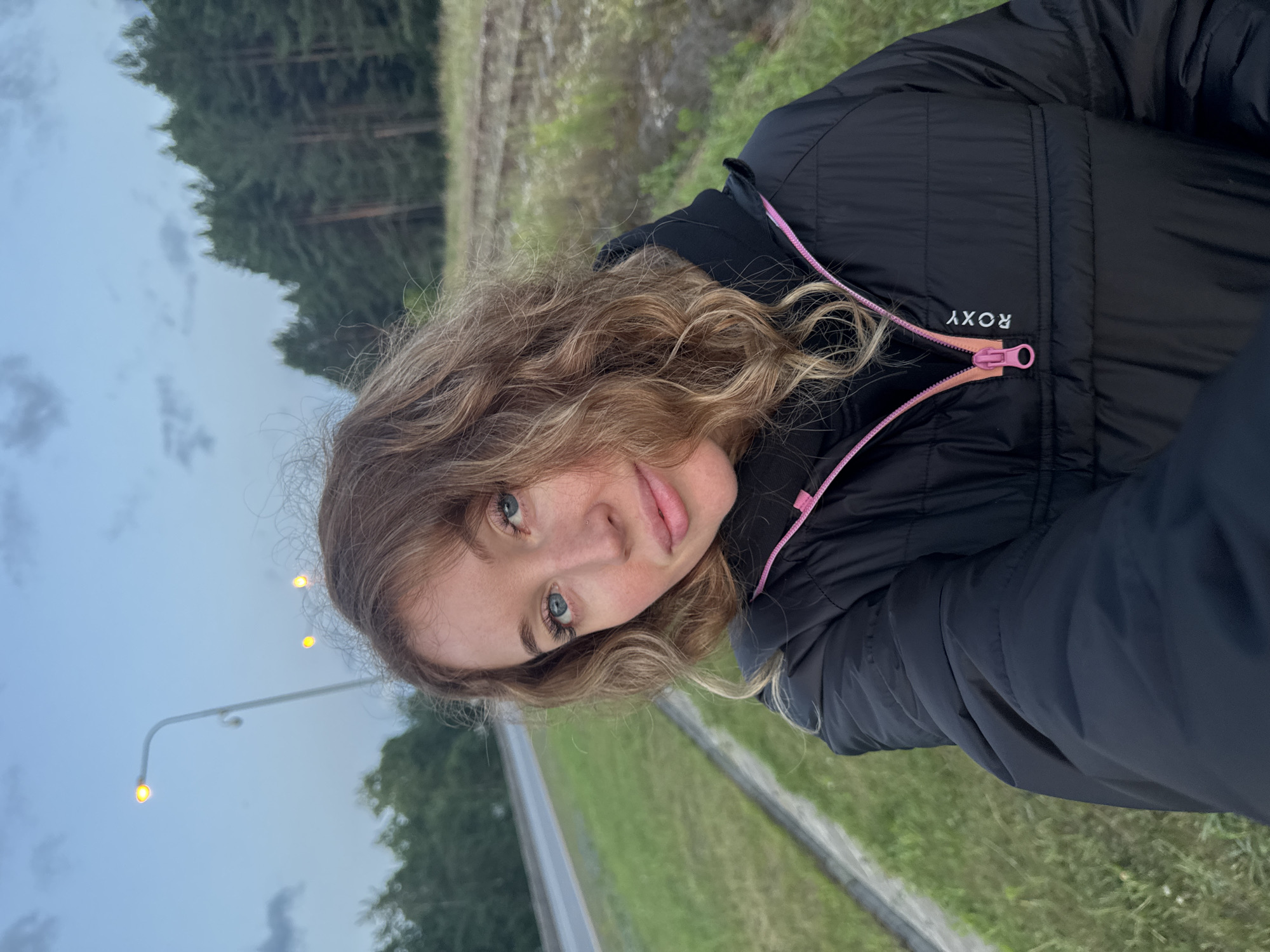 A young woman with wavy blonde hair and blue eyes is taking a selfie outdoors in Chaikovsky, Russia. She is wearing a black quilted jacket with a pink zipper and the brand name "ROXY" printed vertically in white text. The background shows a road with a grassy shoulder, a ditch, a line of pine trees, and a cloudy sky with streetlights visible. The overall lighting suggests it is dusk or early morning.