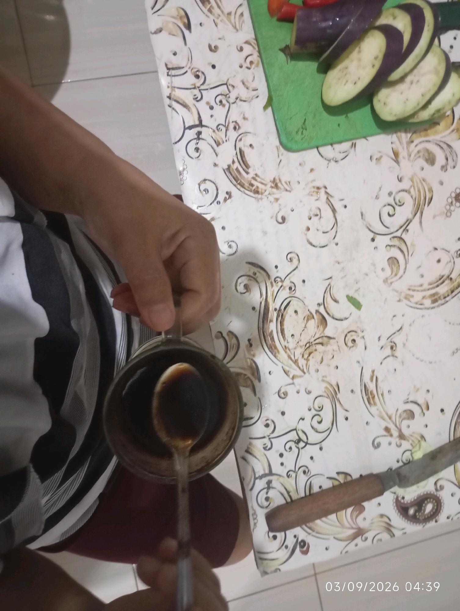 In Klecorejo, Indonesia, a close-up shot reveals a person's hands preparing food on a patterned table. A hand is seen holding a small metal cup filled with dark liquid, possibly coffee, with a spoon resting inside. To the right, a green cutting board holds sliced eggplant and chili peppers, suggesting meal preparation. A knife with a wooden handle lies on the table. The timestamp 03/09/2026 04:39 is visible in the lower right corner of the image.