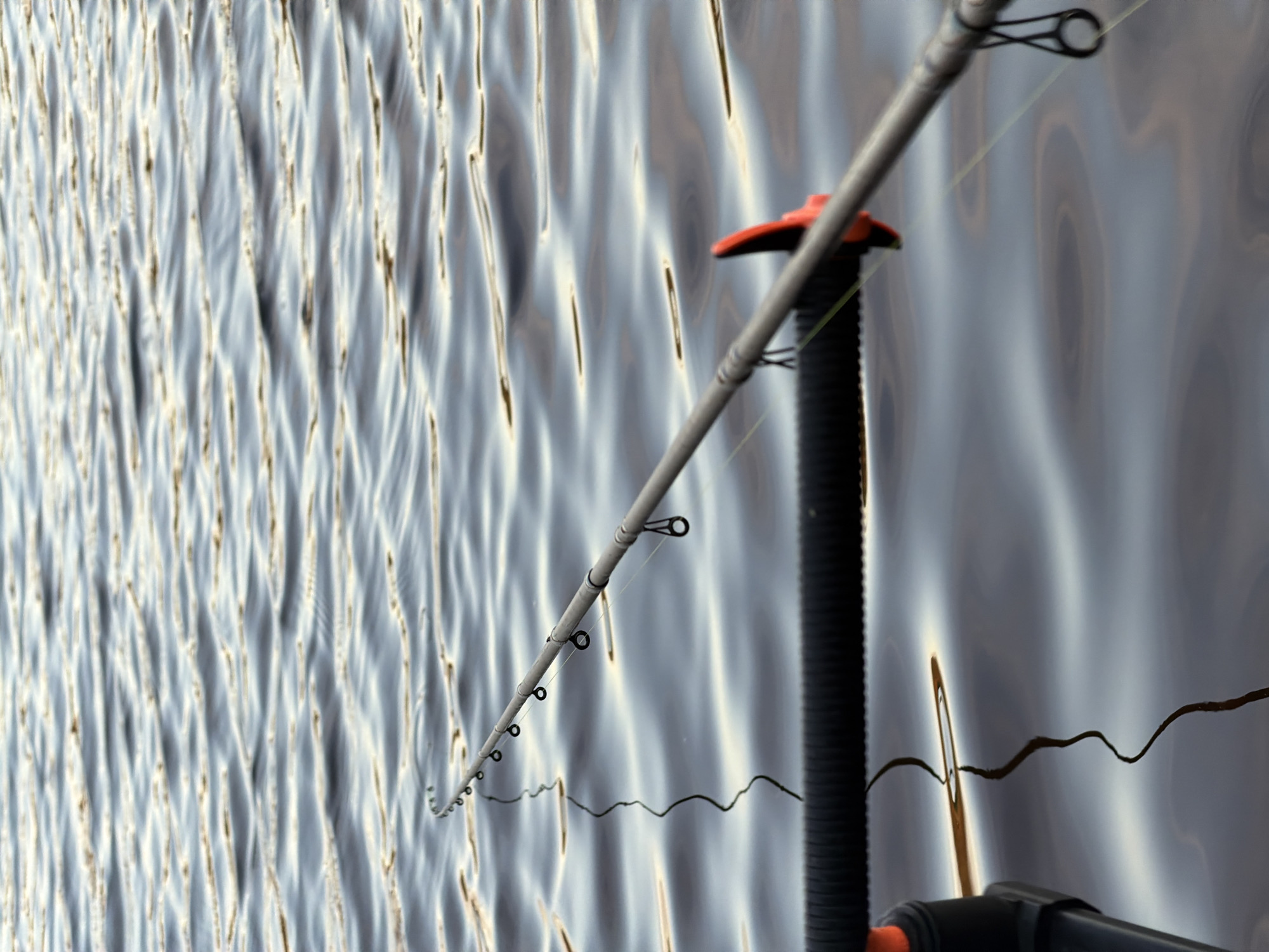 A fishing rod lies horizontally over rippling water, reflecting a muted sky. The rod is gray and equipped with multiple circular guides. A black, ribbed rod holder is visible in the foreground, with an orange accent piece attached to the rod. A dark, wavy line, possibly a fishing line or submerged object, trails into the water from the rod. The scene is set on a body of water, likely a river or lake. There are no people visible in the image. The location is unspecified within the city of Chaikovsky, Russia.