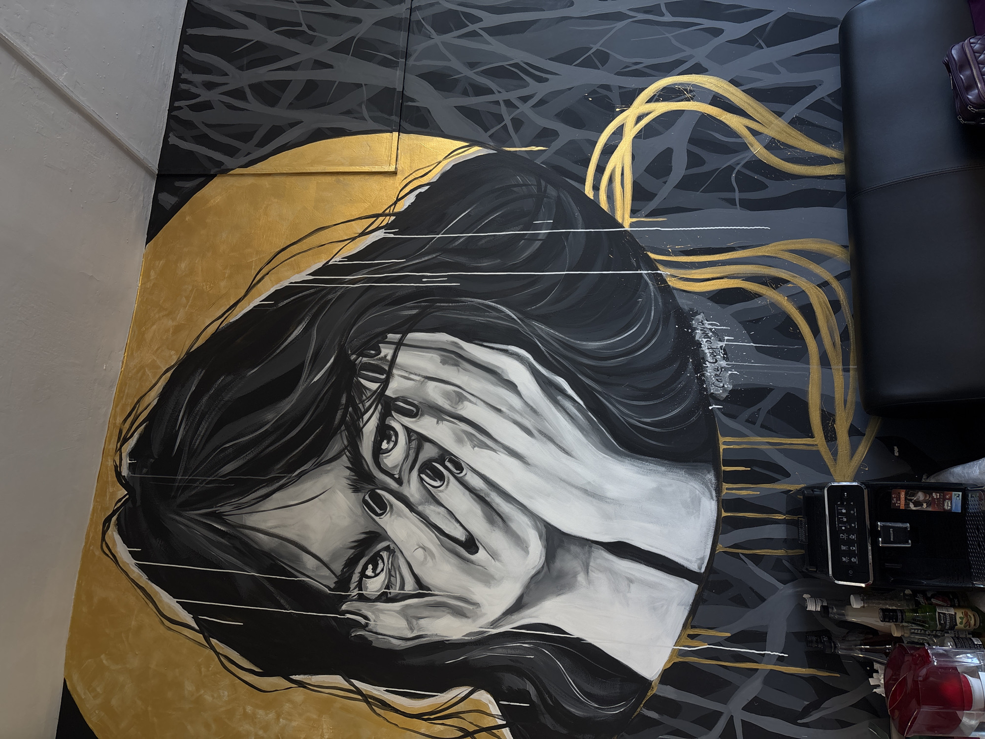 The image captures a low-angle shot of a mural depicting a person covering their face with their hands. The mural is painted on a wall with a section wrapped around to what appears to be a floor. The artwork uses a grayscale palette for the figure, with gold accents in the background around the figure and a gray abstract background of lines. The style appears somewhat surreal or contemporary.
Located near the mural is a black chair and a black Philips coffee machine, along with several bottles. The exact location within the city of Krasnoyarsk, Russia is not discernible from the image itself.