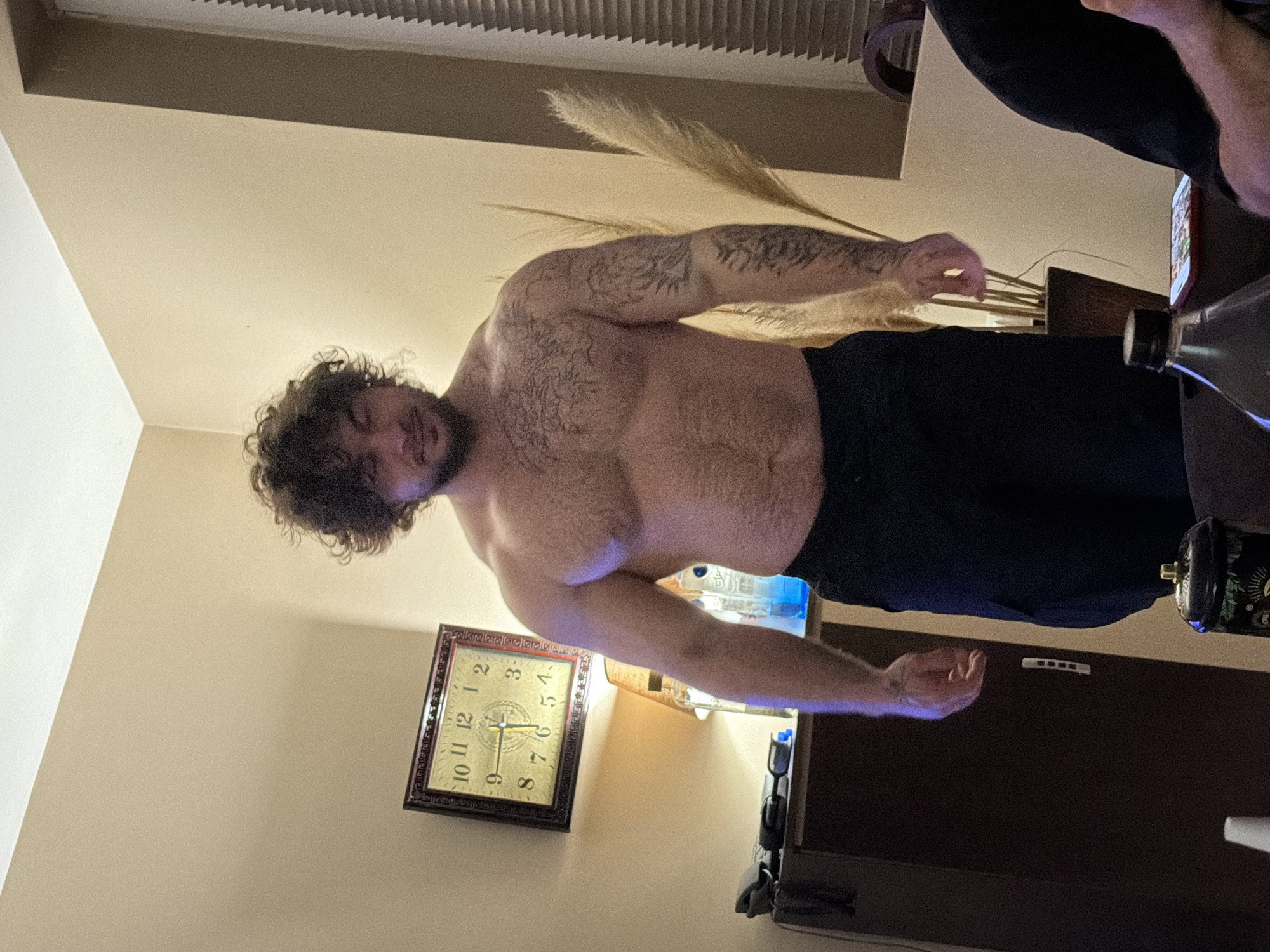 A man with curly hair and tattoos on his arms is standing indoors, shirtless, wearing black pants. A clock is visible on the wall behind him, indicating the time is approximately 6:05. Beside him there is a dark brown piece of furniture and a bottle of water. Location: Unknown, Unknown.