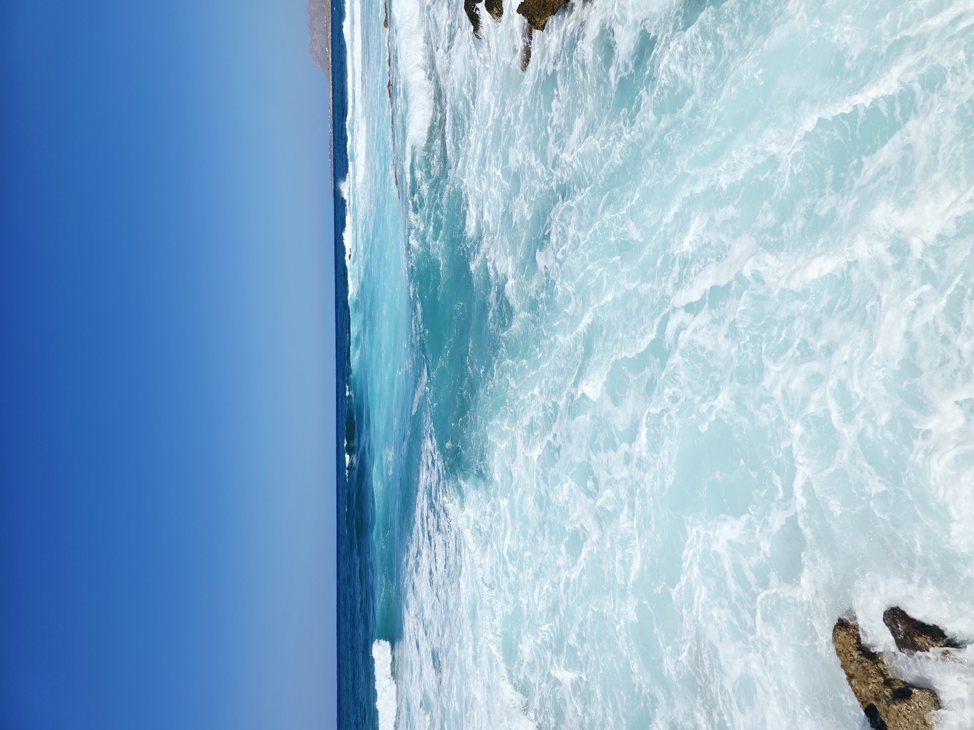The image displays a clear, cloudless blue sky over a turquoise sea with breaking waves. In the foreground, white foamy water churns around scattered rocky outcrops. In the distance, the sea transitions to a deeper blue, and a rocky coastline with sparse vegetation and a few buildings is visible under the bright sky. This scene depicts a coastal area, likely a beach or rocky shore, characteristic of the Chania region in Greece, known for its vibrant blue waters and rugged coastlines. There are no people or specific actions depicted in the image.