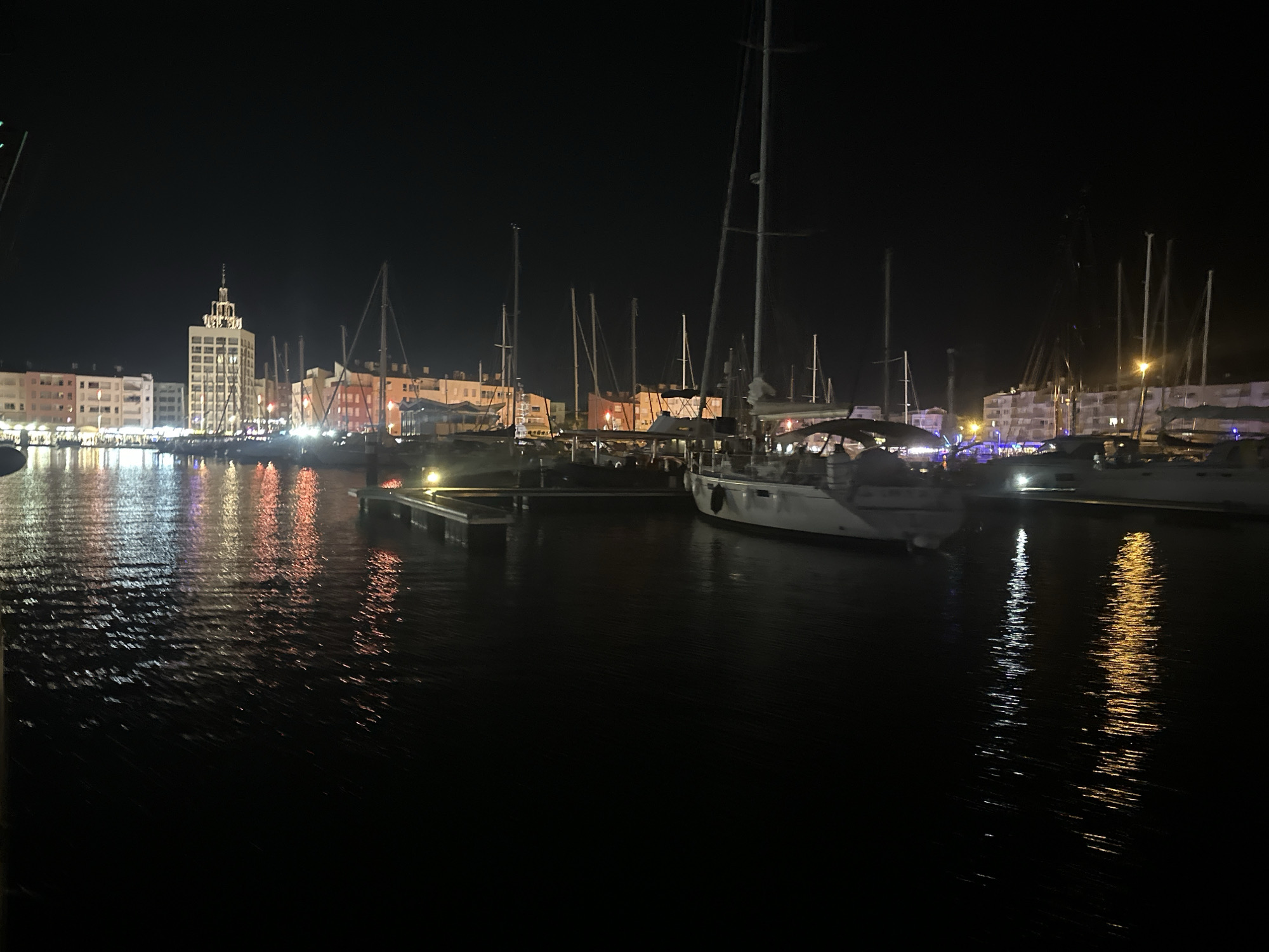 The image captures a nighttime view of the Agde marina in Agde, France, along the Hérault River. The scene is dominated by the reflections of colorful lights from buildings and boats on the dark, rippling water. A tall, illuminated building, likely a significant landmark in Agde, stands out in the background. Several sailboats are moored in the marina, their masts and rigging silhouetted against the night sky and the illuminated cityscape. The water is calm, with the lights creating long, shimmering reflections that stretch across the surface. The overall atmosphere is serene and picturesque, showcasing the beauty of the marina at night.
