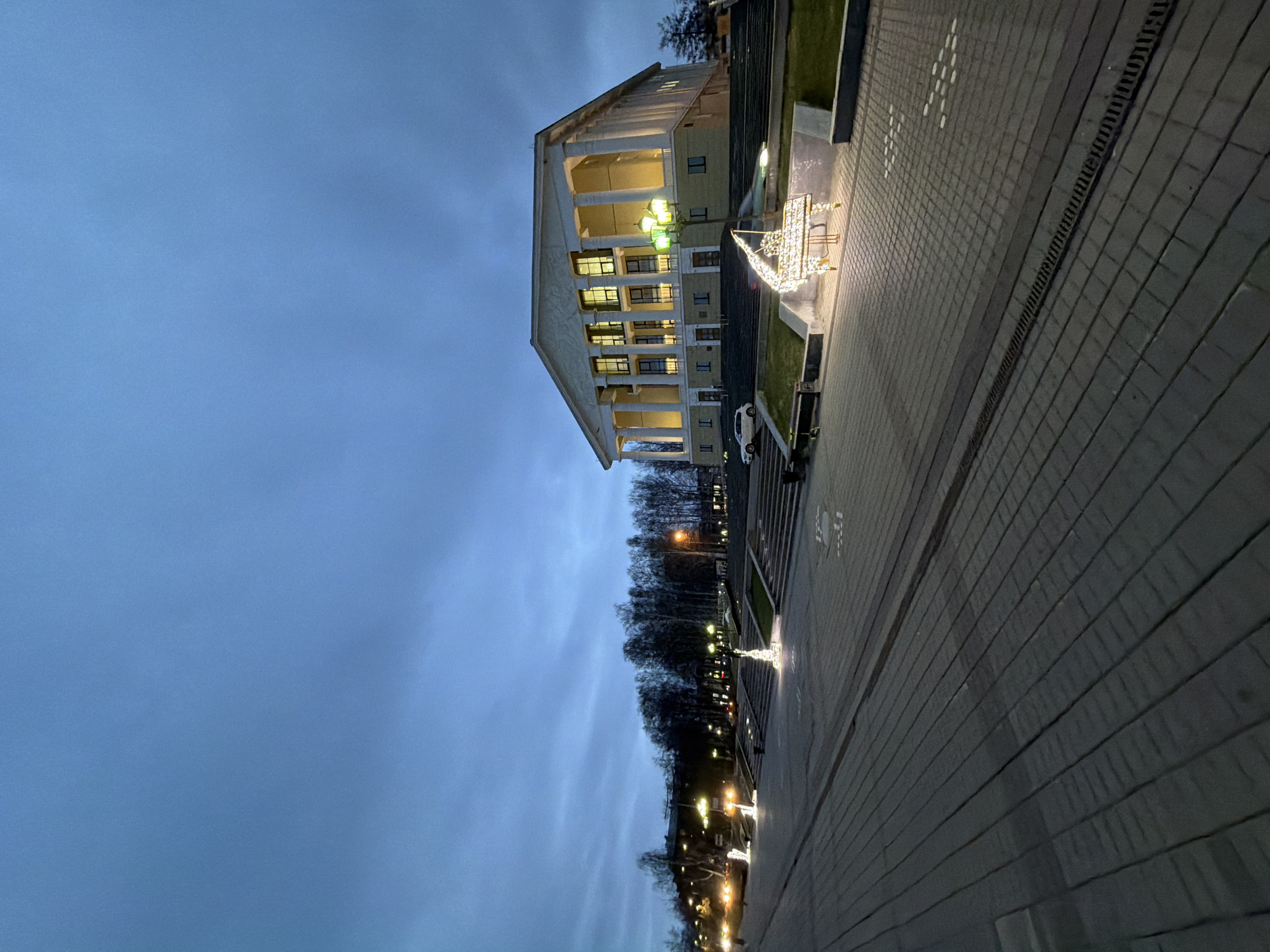 The image shows the building of the Palace of Culture in Votkinsk, Russia, viewed from a slightly low angle. The building is three stories high with a facade featuring columns. Several windows are illuminated from within. A walkway leads to the building, paved with rectangular stones. On the right, there is a line of trees illuminated by lampposts. The sky is a cloudy blue. A decorative light structure resembling a ship or sled is visible on the walkway. A car is parked near the building's entrance.