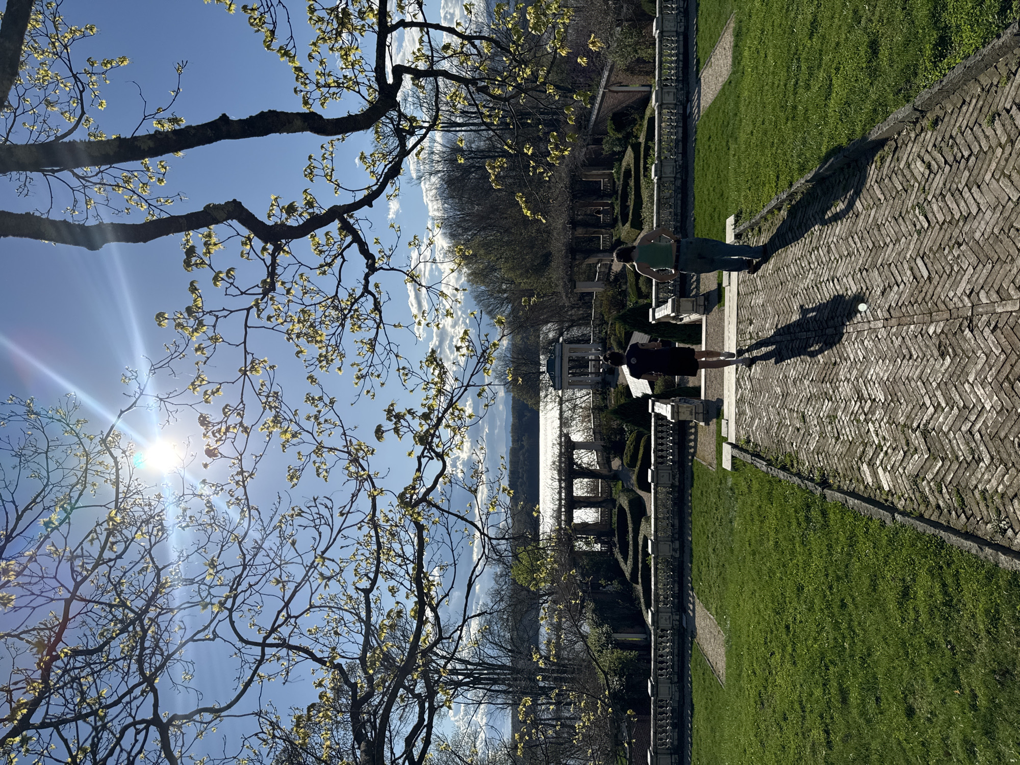 The image shows a scenic view in the Town of Red Hook, United States of America, featuring a garden overlooking a body of water, likely the Hudson River. Two people are standing on a herringbone brick path, casting long shadows. The garden is manicured with neatly trimmed hedges and grass. A decorative balustrade runs along the edge of the garden, defining the space. In the foreground, a tree with budding leaves frames the scene, partially obscuring the bright sun in the upper left corner. The sky is a clear blue with scattered clouds reflecting on the water's surface. In the background, trees line the opposite shore of the river.