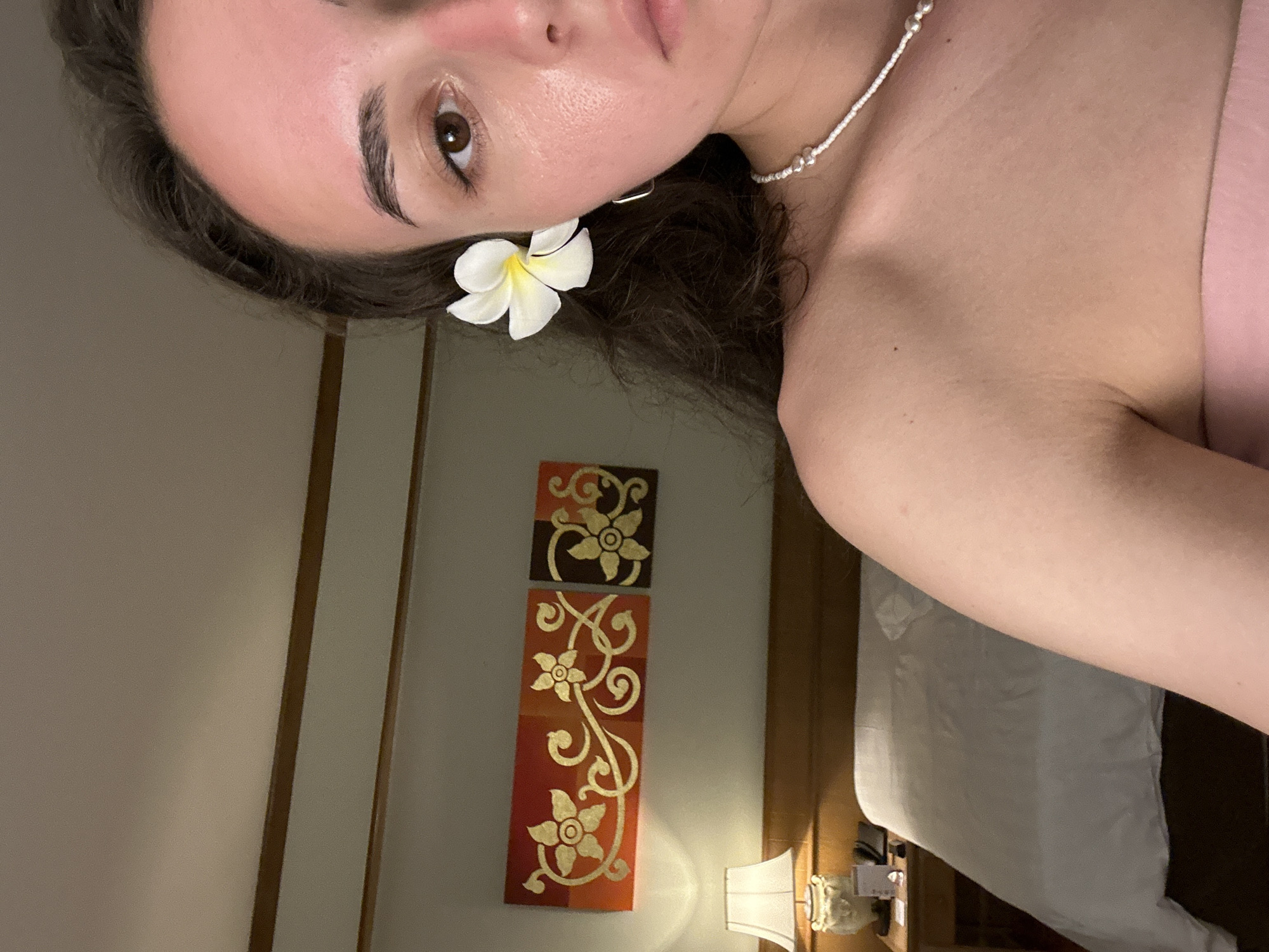 A selfie-style photograph taken indoors in Pattaya City, Thailand, shows a light-skinned female with dark hair. A white flower is tucked behind her ear. She is wearing a beaded necklace. The background includes artwork, a bedside lamp, and a glimpse of a bed.
