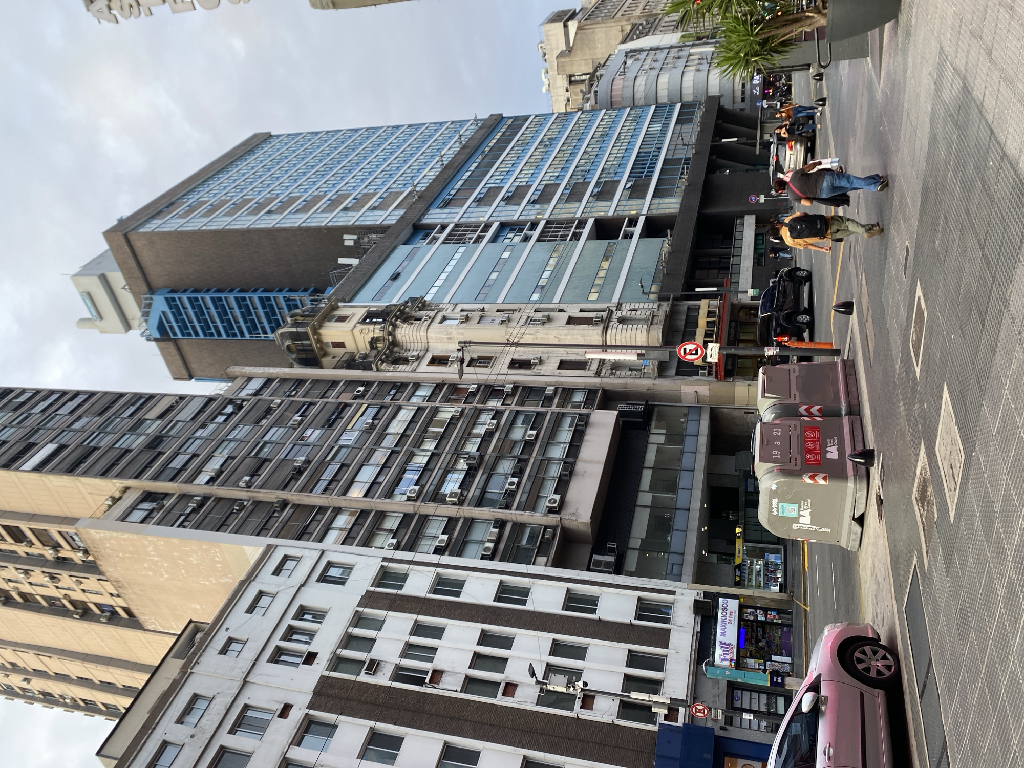 The image depicts an urban street scene in Buenos Aires, Argentina, under an overcast sky. The visual field is dominated by multiple tall buildings of varying architectural styles. On the left, a multi-story building features a white facade with dark brown vertical accents and numerous rectangular windows, some equipped with external air conditioning units. Adjacent to it, a very tall, light-colored building with multiple uniform windows and exterior air conditioning units rises significantly. To the right, a prominent modern high-rise is visible, characterized by dark brown sections and extensive blue-framed windows, with a distinct vertical blue element on one side and a light-colored structure on its roof. An older, ornate building with balconies and a mansard roof is situated below this modern structure.
At street level, a paved sidewalk with textured square panels and metal grates is in the foreground. Two large, grey, rectangular waste containers labeled "19 a 21" and "Buenos Aires Ciudad" are positioned near the curb. A silver sedan is partially visible on the left side of the street, facing away from the viewer. Further down the street, a black pickup truck and other vehicles are present. A red circular sign with a diagonal line through an "E" (indicating no parking/stopping) is mounted on a pole near the sidewalk.
Two pedestrians are walking away from the camera on the sidewalk in the midground: one male wearing a light-colored shirt and a dark backpack, and one female wearing blue jeans and a dark shirt, carrying a bag. Additional pedestrians are visible in the distance. Street lighting fixtures and dark conical bollards are present along the sidewalk. On the left, a "MAXIKIOSCO 24 hrs" storefront with "Fiiu" branding is visible.