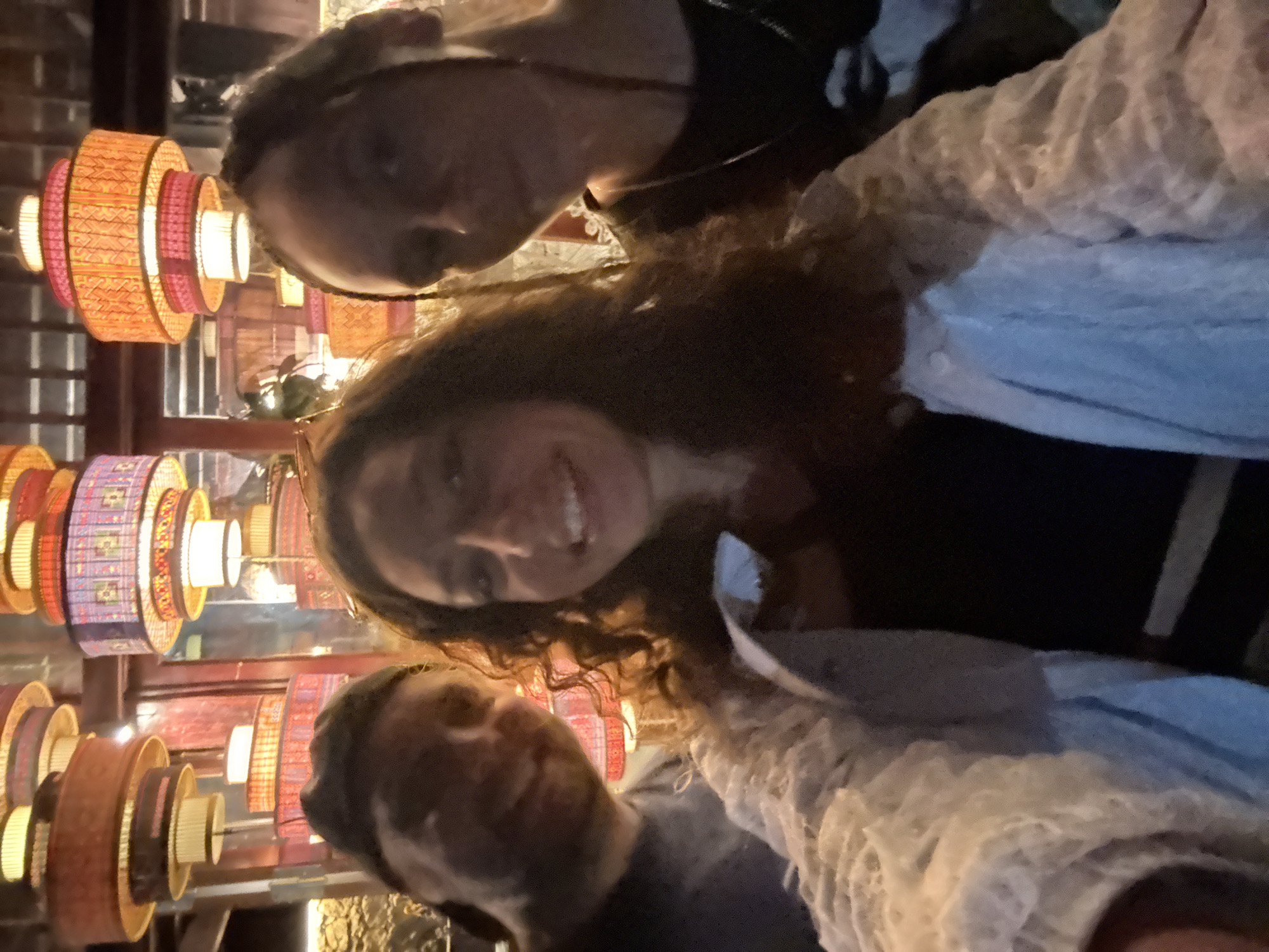 A selfie taken at night in Moc Chau, Vietnam, shows three individuals posing in front of decorative lanterns. The person in the center, a woman with long, curly brown hair, is smiling and wearing a light-colored, textured jacket over a dark top. To her left, a man with a beard and a baseball cap is visible from the shoulders up, also smiling. To her right, another person, whose face is partially obscured, is looking towards the camera. The background is dominated by colorful, tiered lanterns, illuminated from within, creating a warm and inviting atmosphere. The overall scene suggests a casual outing or celebration in a place adorned with traditional or decorative lighting.