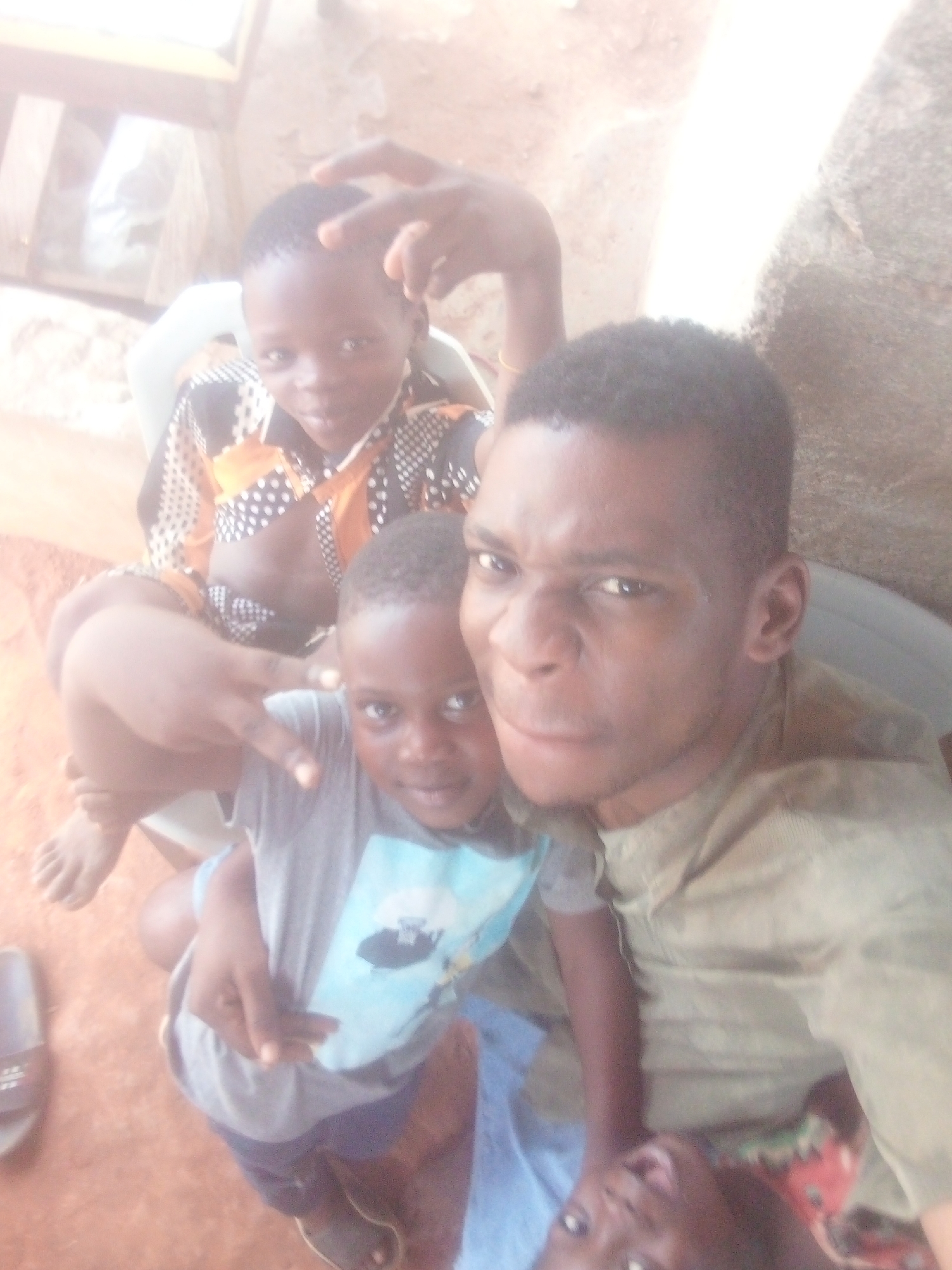 A vibrant, overexposed selfie captures four dark-skinned individuals in a close-up, high-angle shot. The setting appears to be outdoors or in a semi-open space with a reddish-brown dirt ground visible.
At the right, an adult male gazes directly at the camera with a playfully scrunched face, conveying a sense of intensity or mischief. He wears a light-colored, possibly olive green or khaki, collared shirt and has short, dark hair.
To his left, an older child is seated on a white plastic chair. This child, wearing a distinctive black, orange, and white patterned shirt, looks at the camera with a slight smile, raising one hand to make a "peace" or "V" sign near their head, while the other hand gestures similarly. Their bare feet are visible, resting on the ground.
Cradled in front of the adult male is a younger child, also looking at the camera with a neutral expression. This child wears a grey t-shirt with an indistinct light blue graphic and dark shorts. Their hand is also raised, making a "peace" sign. A light blue fabric is draped over their lap.
Partially visible at the very bottom center, another child's eyes and the top of their head peer upwards at the camera, suggesting they are lying down or sitting very low, possibly on a lap or the ground. A colorful fabric, possibly red, green, and white, is near their head.
The image has a bright, almost washed-out quality due to the strong lighting, indicative of daytime. A dark slipper or sandal is on the ground to the far left. The overall mood is casual and intimate, capturing a candid, playful moment among the group. No visible text is present in the image.
