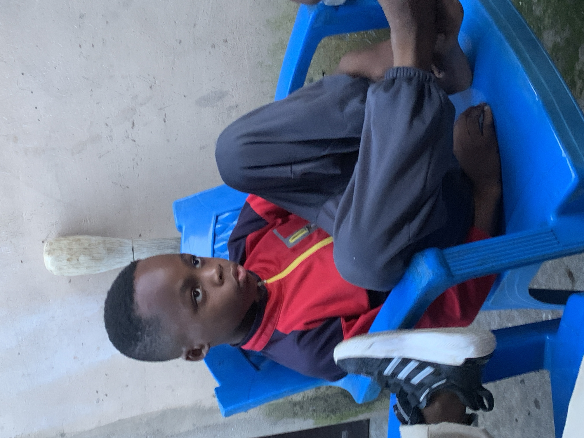 A young Black boy is seated in a bright blue plastic chair, his legs bent and crossed. He is wearing a red and dark blue athletic jacket with yellow trim and dark grey sweatpants. His left foot, clad in a black and white sneaker, rests on the armrest of the chair. The boy looks off to the right with a neutral expression. The background consists of a light-colored concrete wall. The scene appears to be an outdoor setting, possibly a courtyard or backyard in Jos, Nigeria.