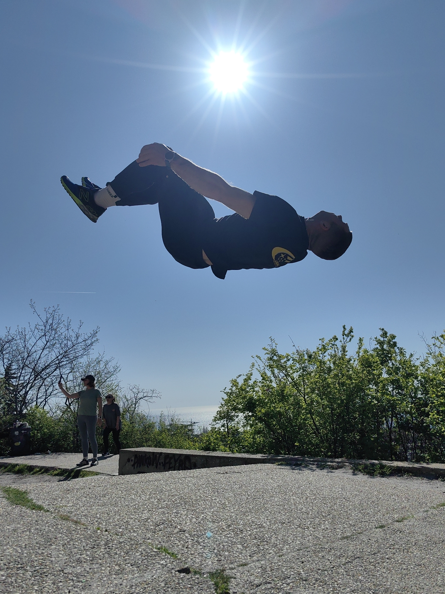 The image captures a man mid-air, performing a backflip with his knees tucked, against a clear blue sky with the sun shining brightly. He is wearing dark-colored pants and a dark t-shirt, and shoes with a lime-green accent and is wearing white socks with “LOSER” written on it in black letters. Below him, the ground is a mix of concrete and a gravelly surface, with greenery consisting of trees and shrubs along the periphery. Two people are visible in the background, on a slightly raised concrete platform. One person is holding a mobile device and appears to be taking a picture or video. A body of water is visible in the distance, suggesting a coastal location. The geographical location is Unknown, Unknown.