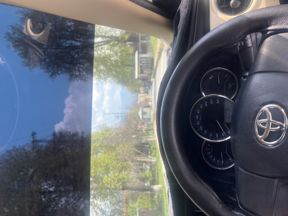 The image presents a forward-facing view from a stationary vehicle, revealing a tree-lined road in Abbottabad, Pakistan. The foreground is dominated by the vehicle's interior: a black, leather-wrapped Toyota steering wheel with a prominent silver logo, positioned directly ahead of a black dashboard covered by a textured black mat. Visible through the steering wheel are the instrument cluster dials, indicating zero speed (0 km/h) and zero RPM, with a digital display showing "13:25" and "41525" (likely odometer). The vehicle's interior trim on the right is beige/tan, with a circular air vent partially visible.
Through the windshield, which has a blue tint at the top, a straight paved road extends into the middle distance. Both sides of the road are lined with mature trees displaying vibrant, light green foliage, indicative of spring or early summer. Further down the road, two small, light-colored guard-hut-like structures are positioned on either side, suggesting a controlled entry or exit point. Beyond these structures, indistinct buildings are visible, and in the far background, hazy blue-grey mountains rise against a partially cloudy blue sky. Tall utility poles and overhead lines are present in the distance. The scene is brightly illuminated by daylight, with visible sun glare on the windshield. No clearly identifiable individuals or specific actions are discernible beyond the implicit presence of the person capturing the image from inside the vehicle. The overall environment suggests an urban or cantonment area typical of Abbottabad, characterized by its greenery and mountainous backdrop.