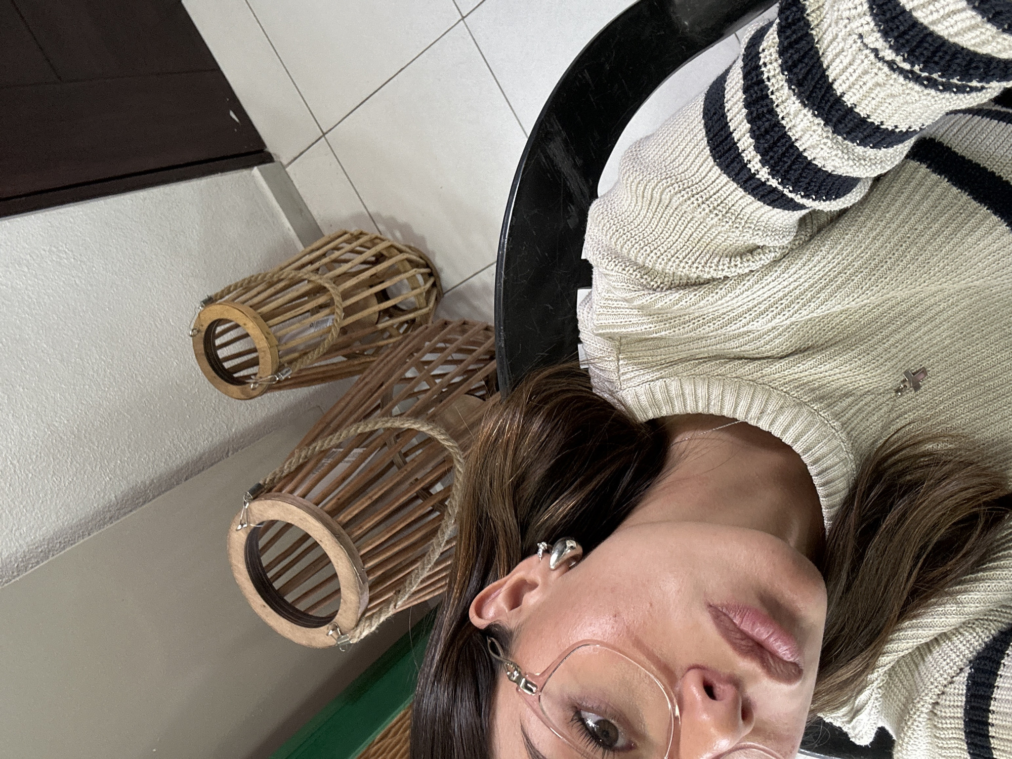 Here's a detailed description of the image:
**Scene:** The image appears to be a selfie taken indoors. The camera is held at an angle, looking slightly downward at the subject.
**Subjects:** The primary subject is a woman wearing glasses and a striped sweater. The sweater features a beige base color with dark horizontal stripes.
**Objects:**
* Two decorative wooden lanterns with rope accents.
* A black table with a glossy surface.
* Neutral-toned floor tiles.
* A dark-colored cabinet or piece of furniture.
* Eyeglasses with a light pink frame
**Environment:** The setting seems to be a modern interior, possibly a home or office. The lighting is generally bright, with light reflecting off the floor tiles and table.
**Location:** Unknown, Unknown. There are no identifiable landmarks or other information to indicate a specific location or city.