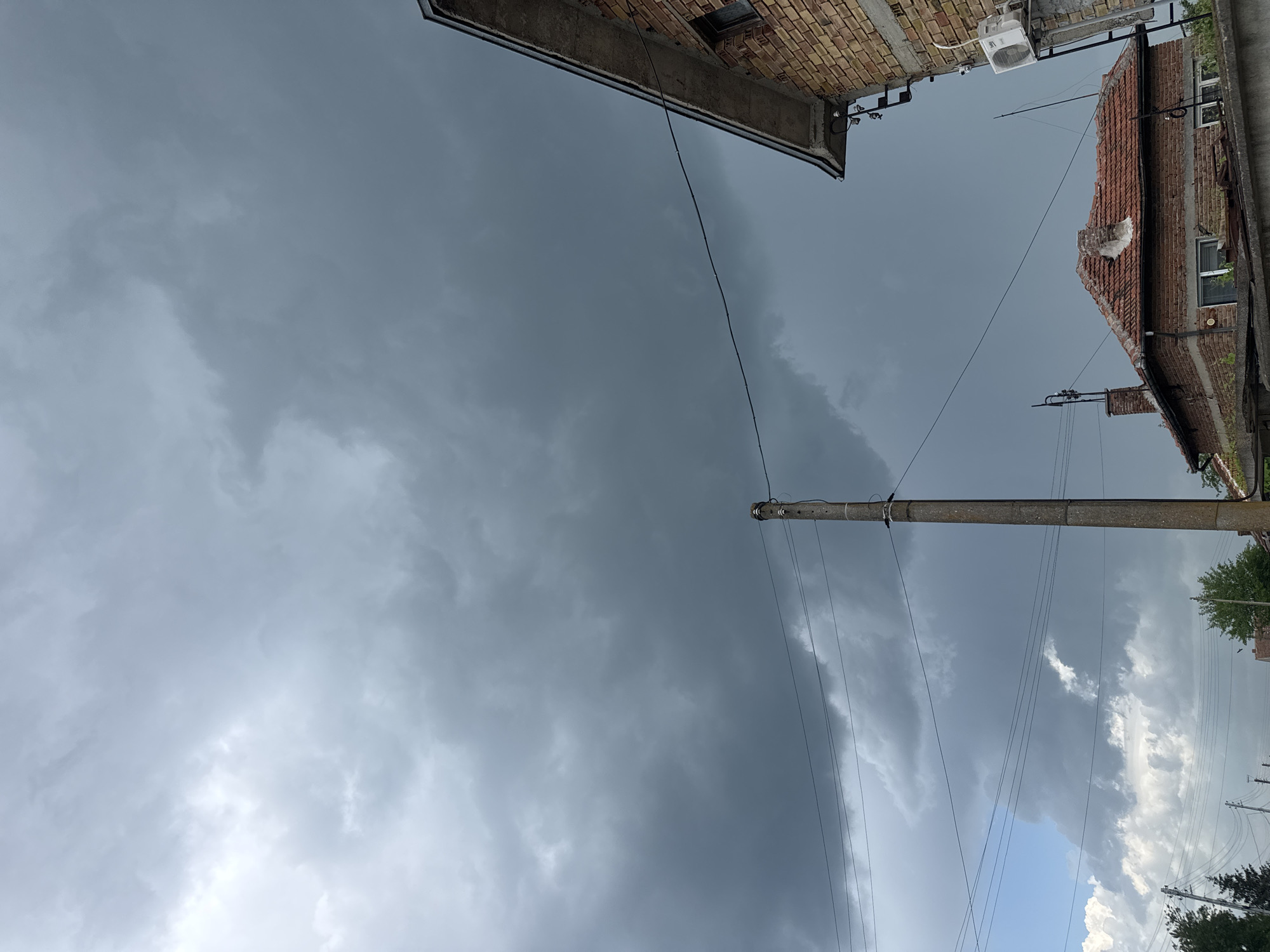 An upward-angled shot captures a sky dominated by dark, ominous clouds, indicative of an approaching storm. Two brick buildings, with details like exposed bricks, windows, and a partial roof, frame the top edges of the scene. A concrete utility pole stands prominently, with several wires stretching across the sky. The lower portion of the image reveals lighter, white clouds, suggesting a contrast in the weather conditions. The image suggests an urban or suburban environment, likely a street corner or alleyway. Based on the provided context, the location is Sarnevo, Bulgaria. No people or animals are visible.