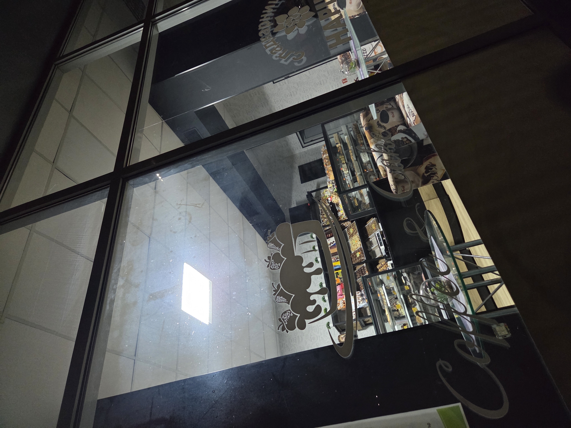 The image depicts a window view reflecting the interior of a building, potentially a café or eatery. Reflected on the window are the words "НАШАТА ПИЦА" (Our Pizza) and "Caffe", suggesting a restaurant. Visible inside are display shelves stocked with goods, indicating the presence of food items. The location is likely in the city of Pleven, Bulgaria, based on the Cyrillic text. The window frame is dark, contrasting with the brighter interior and the reflection of a light source. There are no people directly visible in the reflection.