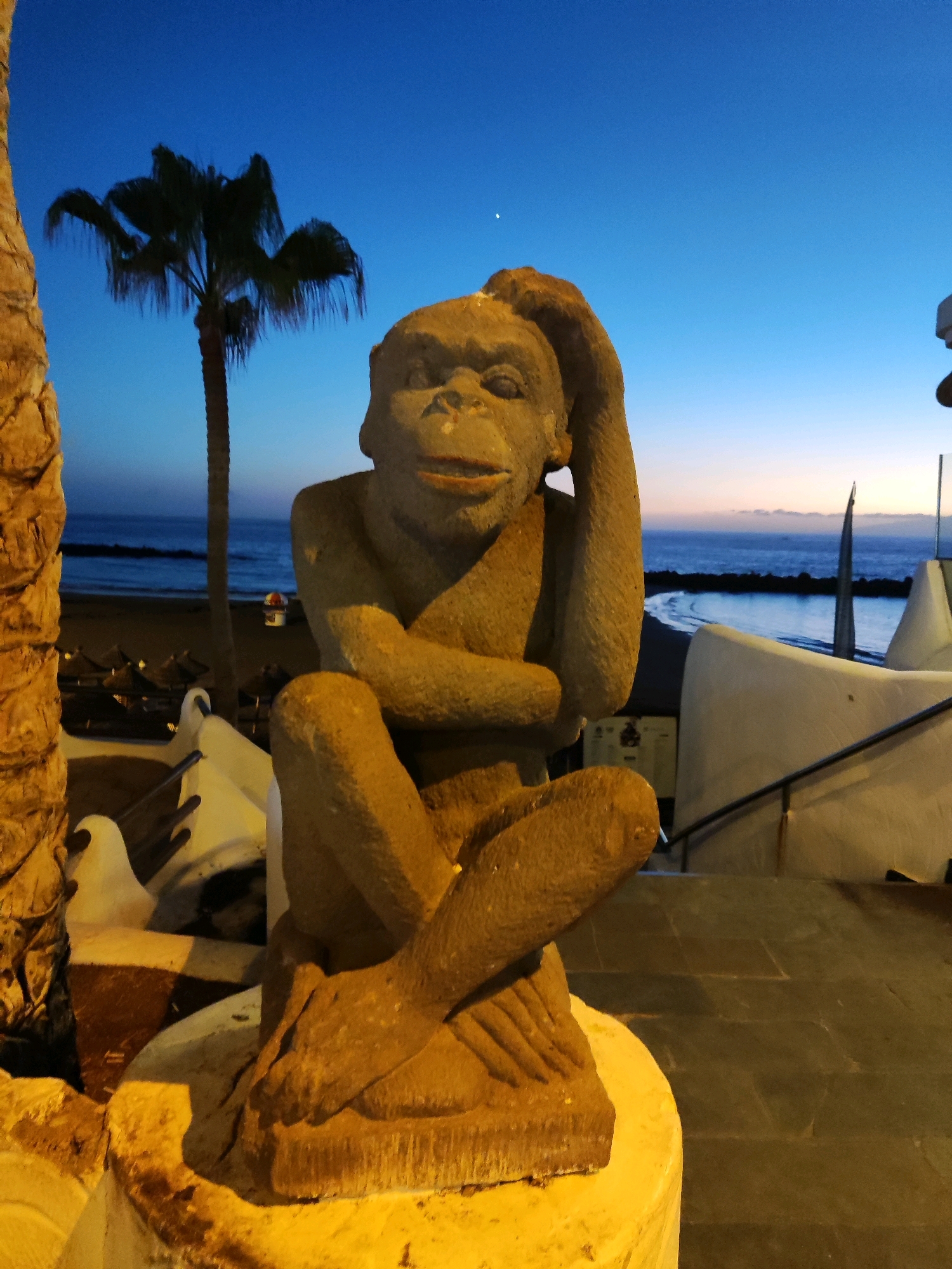 A stone statue of a monkey is positioned in the foreground, seated with one hand behind its head and the other resting on its lap. The statue is on a circular, yellow base. Behind the statue, a palm tree is visible against a twilight sky with a gradient of blue to a faint orange near the horizon. The ocean is visible in the background, with gentle waves and a dark shoreline. A few thatched umbrellas and loungers are sparsely dotted along the beach. To the right of the statue, modern white structures with curved elements and dark railings are present, suggesting an outdoor seating or dining area of a resort or establishment. The overall scene depicts an outdoor setting at dusk, likely a coastal resort area. This statue is located in the town of Adeje, Spain, a popular tourist destination on the island of Tenerife, known for its beaches and resorts.
