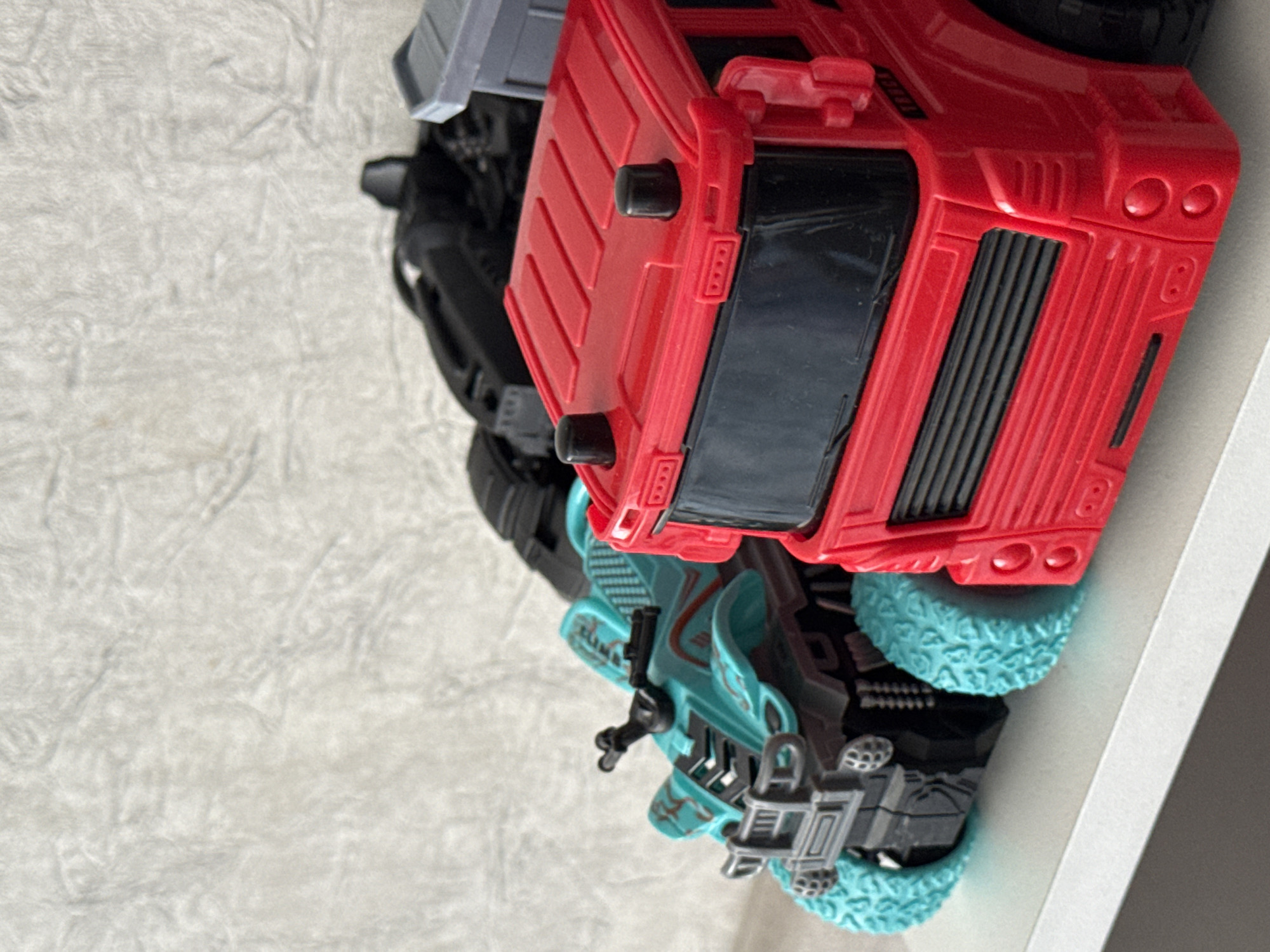 The image displays a close-up, eye-level shot of toy vehicles arranged on a white surface against a textured, off-white background. On the right is a red toy truck, featuring black headlights, a grille, and a windshield. Two black circular lights are present on its roof. To the left of the truck is a teal-colored toy all-terrain vehicle (ATV) with black accents and bright blue, textured wheels. Behind the ATV, a black toy vehicle with a complex structure is partially visible. The scene appears to be indoors, likely on a shelf or table. The location is specified as Yarominski Selski Sovet, Belarus, though this detail is not visually inferable from the image itself.