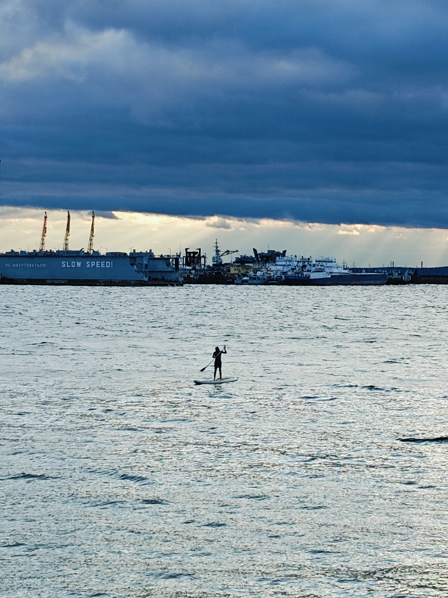 In the city of Chaikovsky, Russia, on a body of water, a solitary figure is paddleboarding. The individual is silhouetted against the water, holding a paddle upright. In the background, a large industrial barge with the text "SLOW SPEED!" is visible, along with cranes and other docked vessels, indicating a port or industrial area. The sky above is filled with dark, dramatic clouds, with rays of light breaking through in the distance. The water is choppy, reflecting the light and the overcast sky.