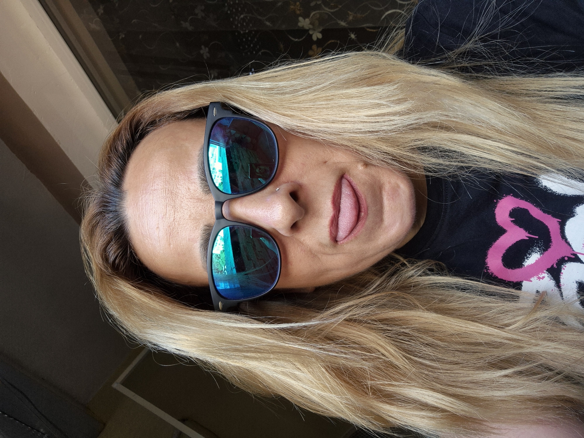A woman with blonde hair and sunglasses sticks her tongue out at the camera. She is wearing a black t-shirt with a pink heart graphic. The background appears to be indoors with a window or curtain behind her.