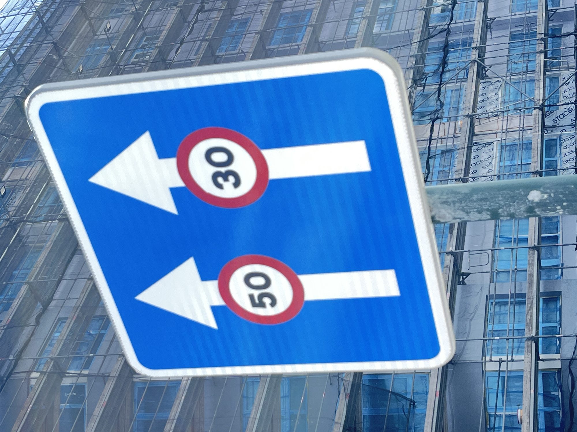 The image depicts a blue and white road sign with two arrows pointing left, each accompanied by a red circle containing a number. The top arrow has a "30" inside its circle, while the bottom arrow has a "50". The sign's background includes a building façade undergoing construction, partially obscured by scaffolding and protective sheeting. Words can be partially read in the sheeting such as "ACBO" and "STACBOND". The photo appears to be taken outdoors, in daylight. The sign appears to indicate speed limits for traffic going in specific directions. Although specific locations is not available from provided data, the OCR data from the building sheeting mentions the name "STACBOND" which has a known association with the city of A Coruña, Spain.