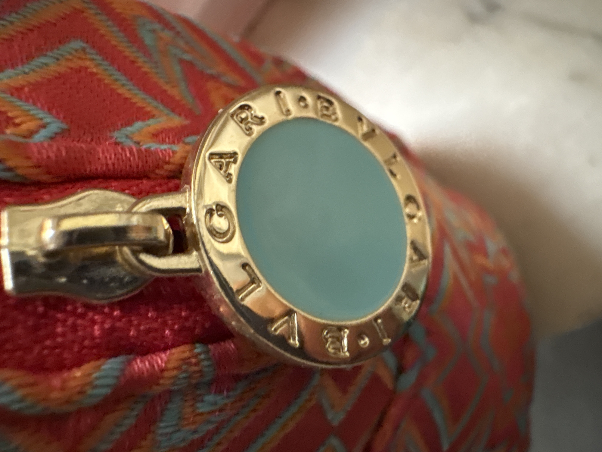 The image shows a close-up of a gold-tone zipper pull on a red, orange, and blue patterned fabric. The zipper pull is oval-shaped and features the embossed letters "BVLGARI" around a light blue circular inlay. The fabric appears to be part of a small pouch or bag, with a zigzag pattern. The background is out of focus, showing a blurred white surface that could be a mannequin or a table. There are no people or other distinguishing environmental features visible. The image does not provide any information about a location in Rome, Italy.