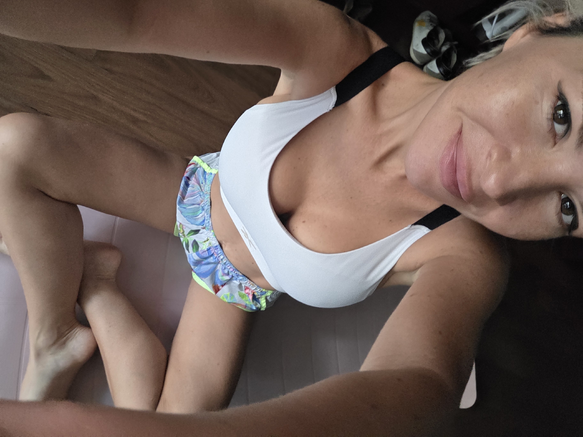 The media file is a selfie taken from a high-angle perspective, featuring a woman in athletic wear. She is kneeling on what appears to be a yoga mat, with a wooden floor visible in the background. The woman has blonde hair and is looking towards the camera with a slight smile. She is wearing a white sports bra with black straps and colorful patterned shorts. Her legs are bent, and her arms are positioned in a way that suggests she is taking the photo herself. In the background, a pair of athletic shoes is partially visible. The lighting suggests it is daytime, possibly indoors. There is no visible text in the image. The setting appears to be a home or gym environment. The woman's expression is relaxed and casual, indicating a moment of personal activity or self-documentation.