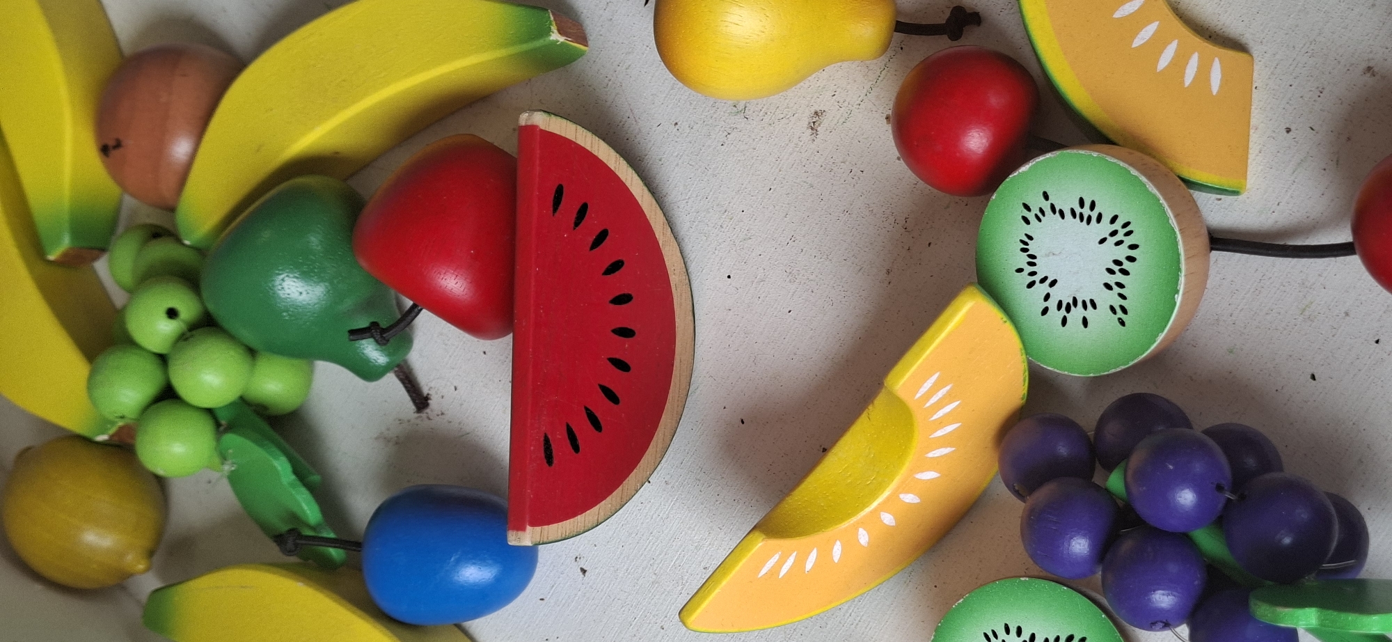 This is a close-up, eye-level, still photograph of a collection of brightly colored wooden toy fruits arranged on a textured, light-colored surface, possibly a table or a floor. The lighting is diffused, casting soft shadows and highlighting the glossy finish of the toys. The scene is devoid of any people or discernible background elements that could pinpoint a specific location within Naantali, Finland, or elsewhere.
The fruits include:
- A large slice of watermelon, red with black seeds.
- Two bright yellow bananas, one positioned horizontally and the other diagonally.
- A cluster of green grapes, each individual grape a small, round sphere.
- A slice of orange-colored fruit, possibly cantaloupe or honeydew, with white markings.
- Two slices of kiwi fruit, green with black seeds arranged in a circular pattern.
- A red apple or tomato-shaped fruit.
- A blue spherical fruit.
- A yellow pear-shaped fruit.
- A brown spherical fruit, possibly a wooden ball.
- Two green leaf-shaped pieces, one attached to the grapes and the other to a blue fruit.
- A red cherry-shaped fruit.
The toys are positioned in a somewhat scattered arrangement, with some overlapping. The overall impression is that of a playful, organized display of children's playthings.