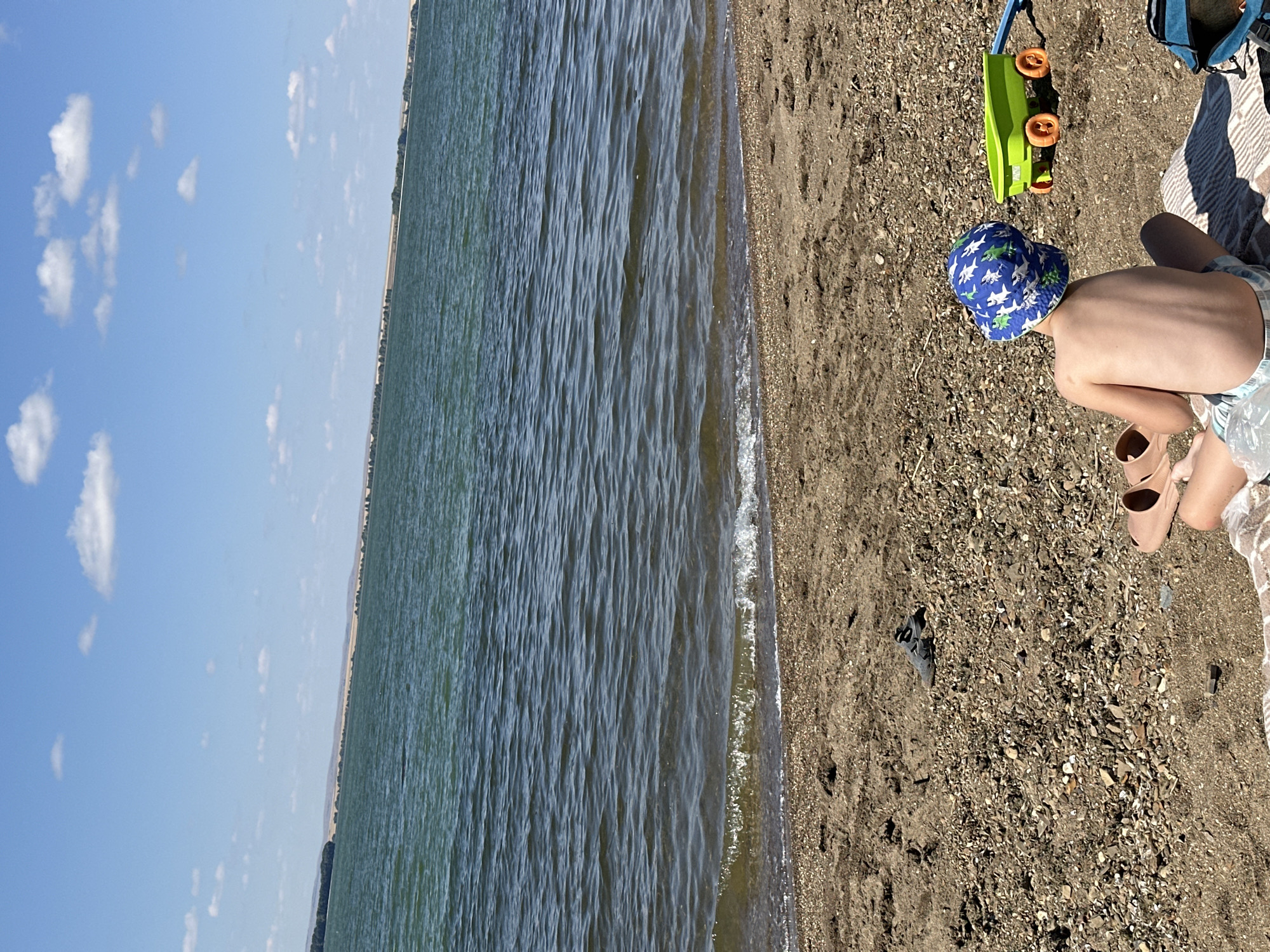 A child wearing a blue and green shark-patterned sun hat and light blue swim trunks sits facing away from the camera on a beach blanket near the edge of a large body of water. The child is shirtless. To the right of the child is a green plastic toy wagon with orange wheels. To the left of the child, also on the beach, are two pairs of sandals, one a darker color and one a lighter, fleshy tone. Further to the left, a single dark shoe rests on the pebbly sand. The water is a deep blue-green, with small waves lapping at the shore. In the background, a distant treeline and rolling hills are visible under a bright blue sky with scattered white clouds. The image appears to be taken on a sunny day in Energetik, Russia, likely near the Energetik Reservoir.