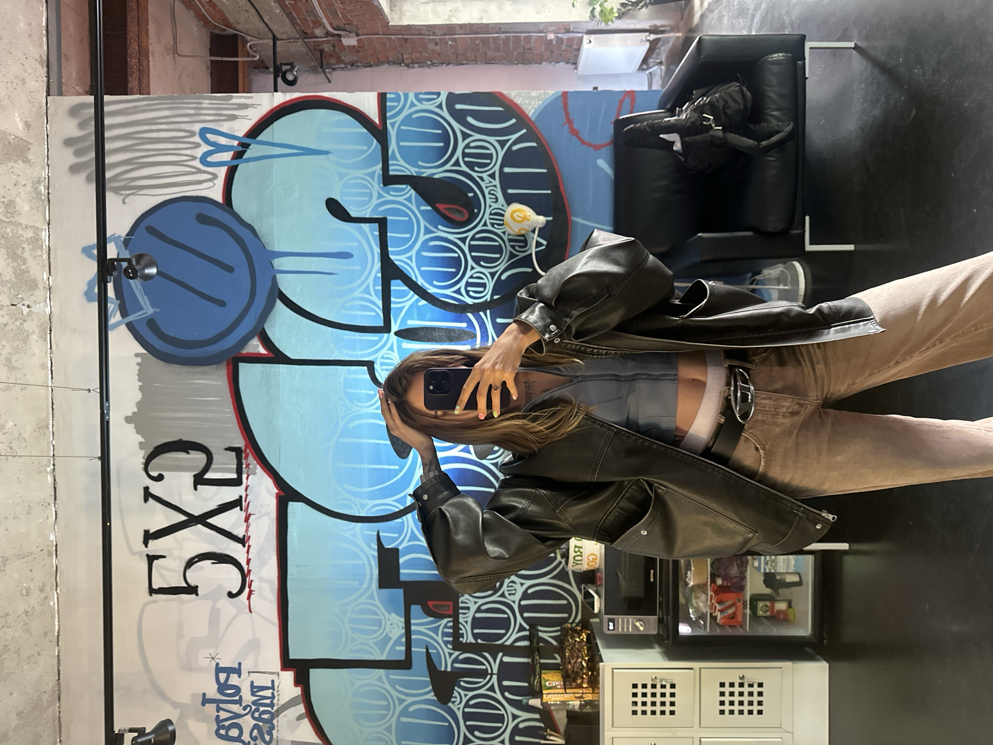 This is a vertical, full-body mirror selfie of a woman. She is standing in front of a graffiti-covered wall, with a black leather jacket over a grey top, and beige pants. She is holding a black smartphone up to take the picture, with her other hand touching her hair. The graffiti on the wall is large and colorful, with blue and white swirls and red accents. There are also some grey scribbles at the top left of the wall.
In the background, there is a black leather couch with a black backpack on it. To the right of the couch, there is a dark floor and a small black mini-fridge with various items inside, including what looks like bottles of water and snacks. Above the mini-fridge, there are some white drawers with square patterns on them.
The setting appears to be an indoor space, possibly a studio or a room with exposed brick on the ceiling, suggesting an urban or industrial environment. The lighting is bright, indicating it's daytime. There's no indication of weather. The overall mood seems casual and self-aware, as typical of a mirror selfie.
Visible text on the wall includes "5XG" and "loyl 26M" in black lettering, along with other illegible graffiti. On the mini-fridge, some labels are visible, but the text is too small to read clearly.
