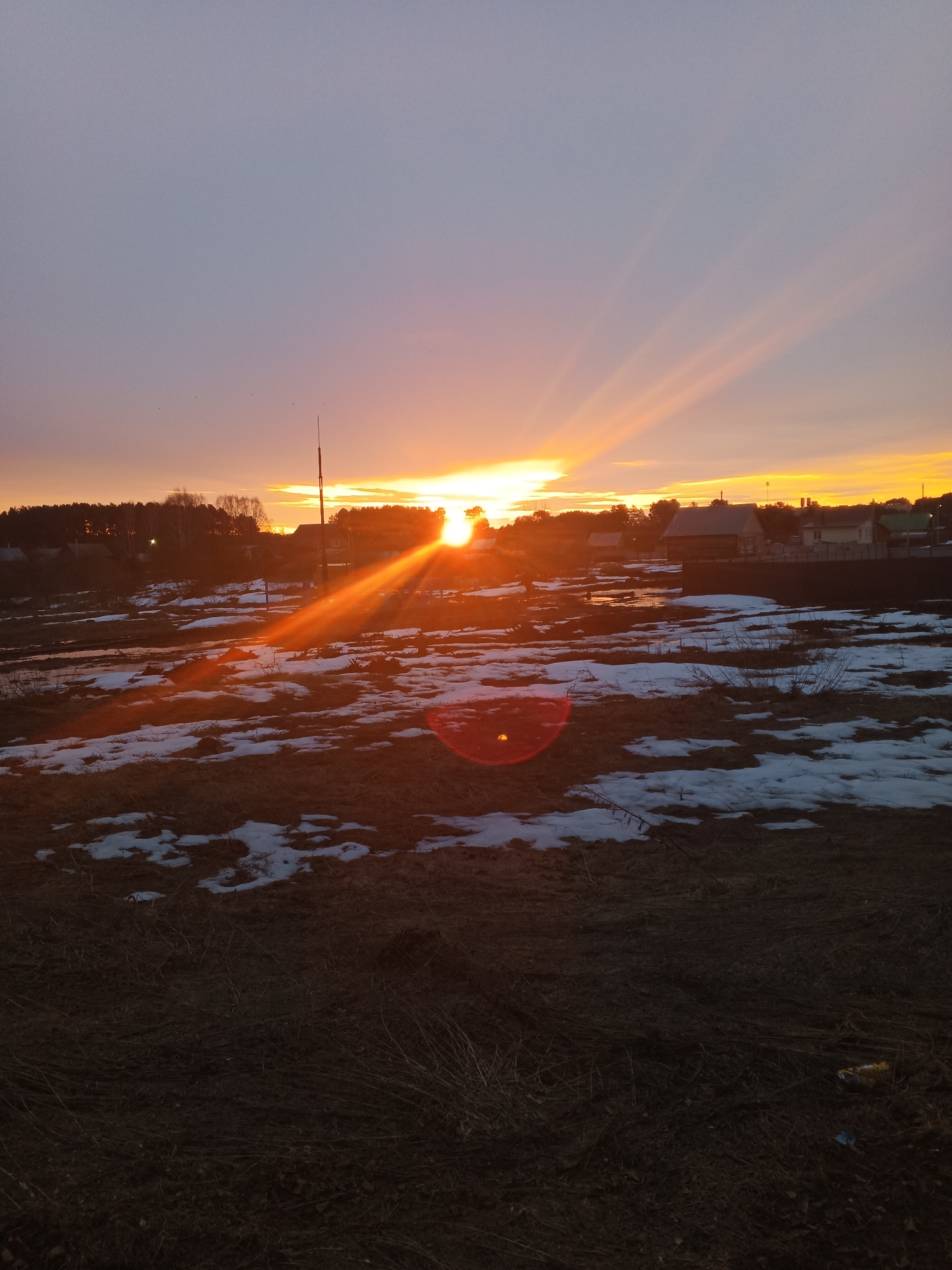 The image shows a landscape at sunset, likely in the vicinity of Rusalevka, Russia. The foreground is dominated by dark, grassy terrain with patches of melting snow. In the middle ground, there are bodies of water reflecting the orange and yellow hues of the setting sun. A line of trees and buildings is silhouetted against the bright horizon. The sun's rays are visible, creating streaks of light in the sky and reflections on the snow and water. A tall, slender structure, possibly a utility pole, is visible among the buildings. There are no people visible in the image. The overall scene conveys a sense of rural landscape in a transitional season.