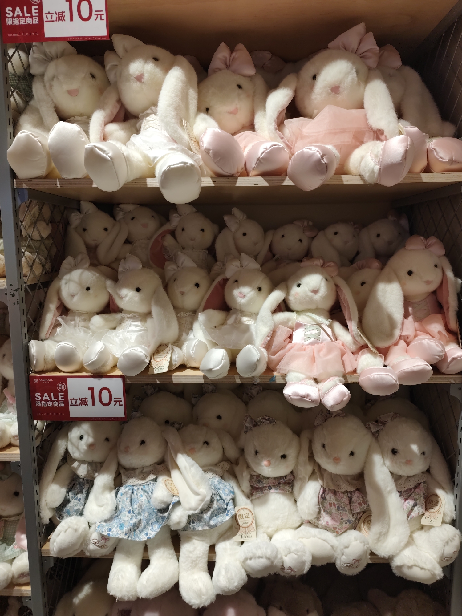 The image depicts several shelves filled with white plush bunny stuffed animals in a retail setting. The bunnies are adorned with bows and various outfits, including pink tutus and blue floral dresses. Some of the bunnies have tags, and a red and white "SALE" sign with Chinese characters and the number "10" is visible on the top shelf. The overall environment suggests a toy store or a department store display. The question about the location in Daqing, China cannot be answered from the provided image.