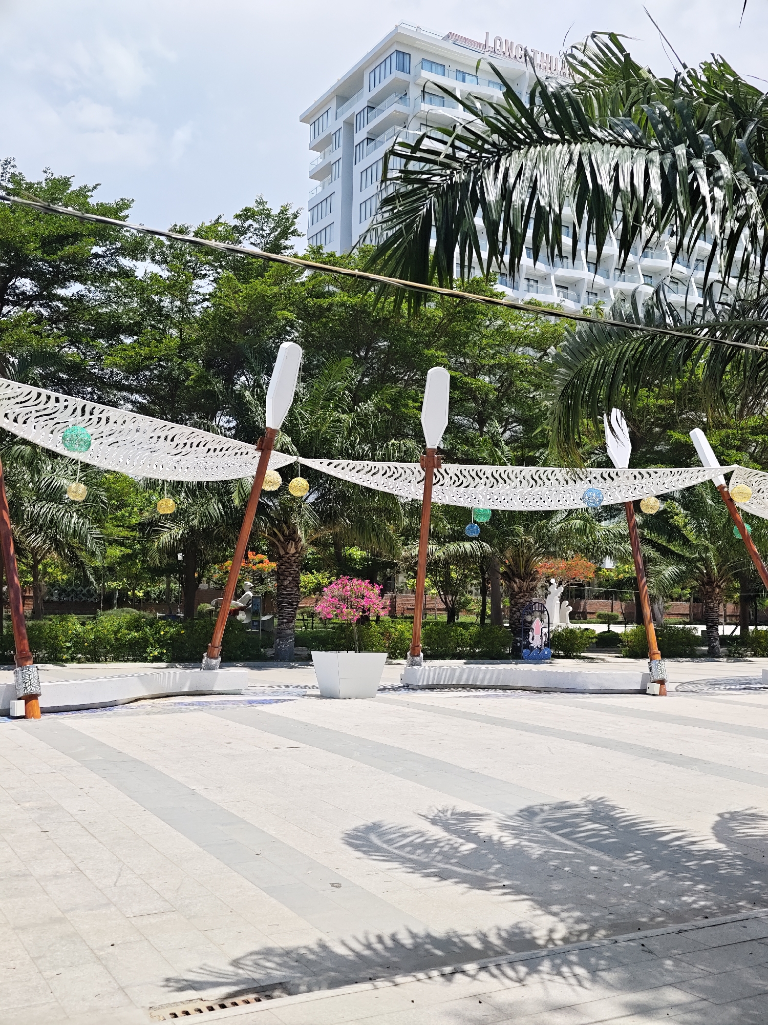 An outdoor area in Phan Rang – Tháp Chàm, Vietnam, features a paved walkway with a striped pattern. In the background, a multi-story white building with balconies is visible, bearing the signage "LONG THUẬN". In the mid-ground, a decorative canopy is supported by wooden poles, adorned with white latticework and hanging spherical ornaments in green and yellow. There are also two tall, white, paddle-like structures. A white planter contains a small tree with pink blossoms. Further back, a sculpture of a tentacled creature can be seen. Lush green trees and palm trees surround the area. The ground is shaded by palm fronds, casting distinctive shadows. A storm drain is visible in the foreground.
